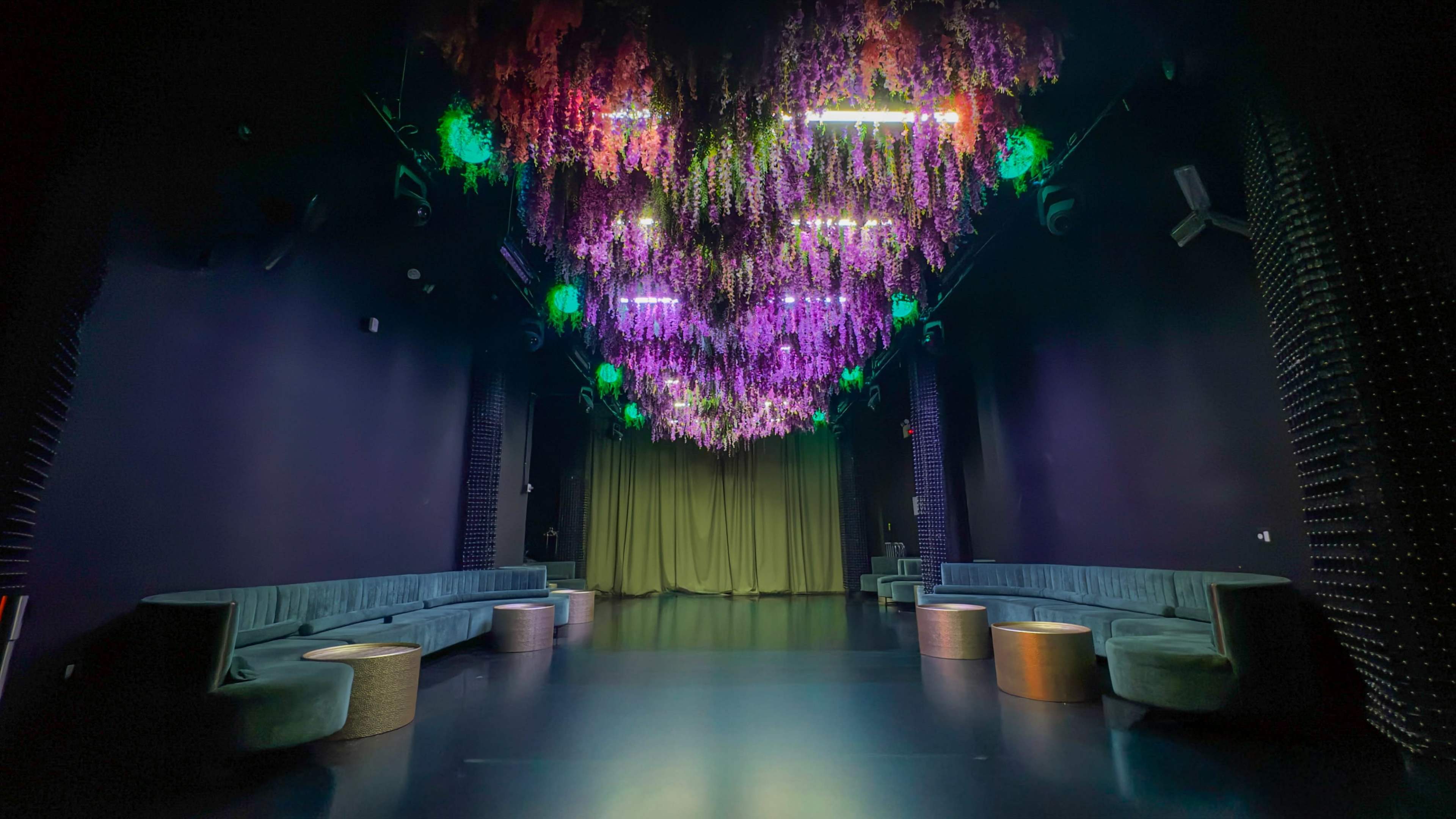 The image shows a dimly lit event space featuring a vibrant floral installation hanging from the ceiling, with seating arrangements along the walls.