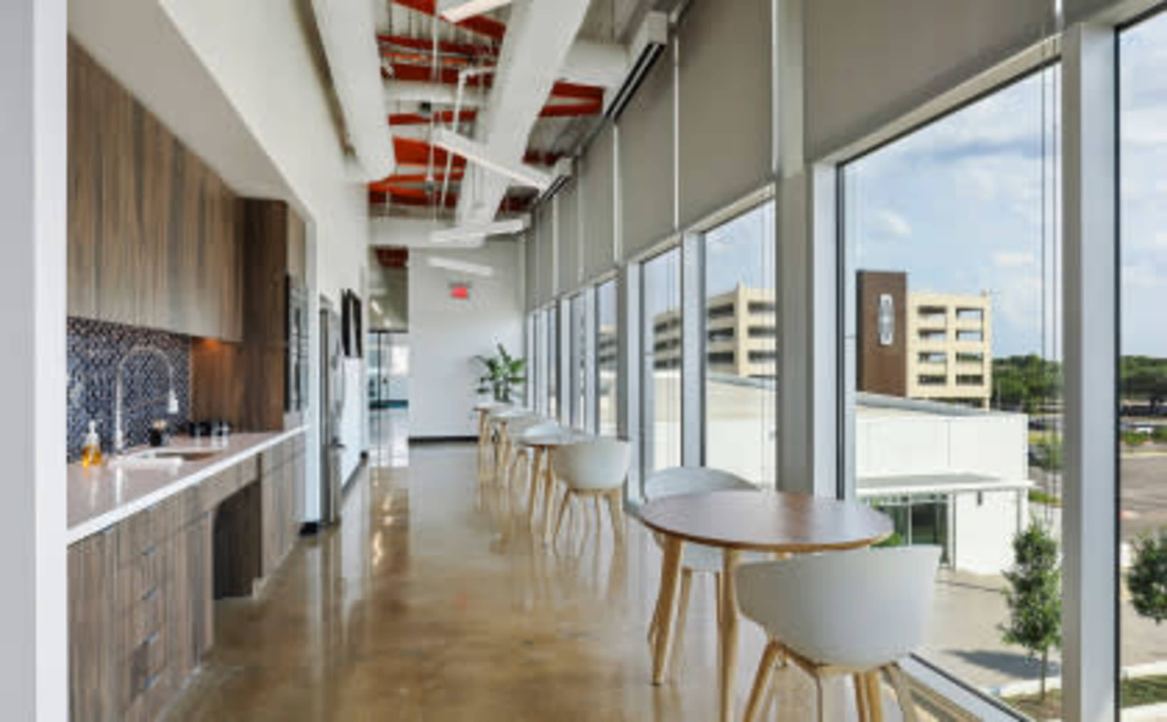 The image shows a modern office kitchen area with large windows, wooden cabinetry, and round tables overlooking a parking lot.