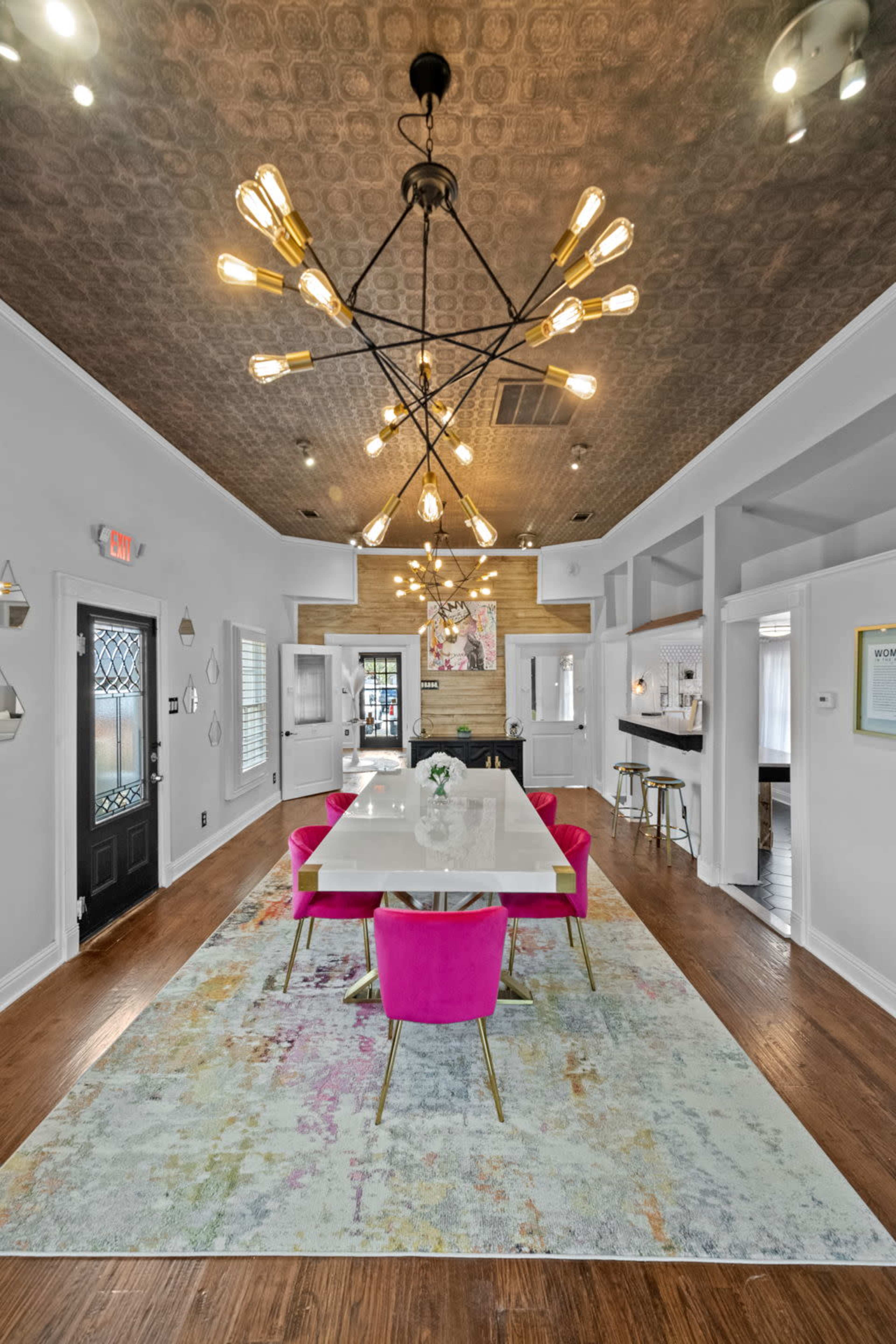 A modern dining area featuring a white table surrounded by pink chairs, with a decorative light fixture and a patterned rug on a wooden floor.