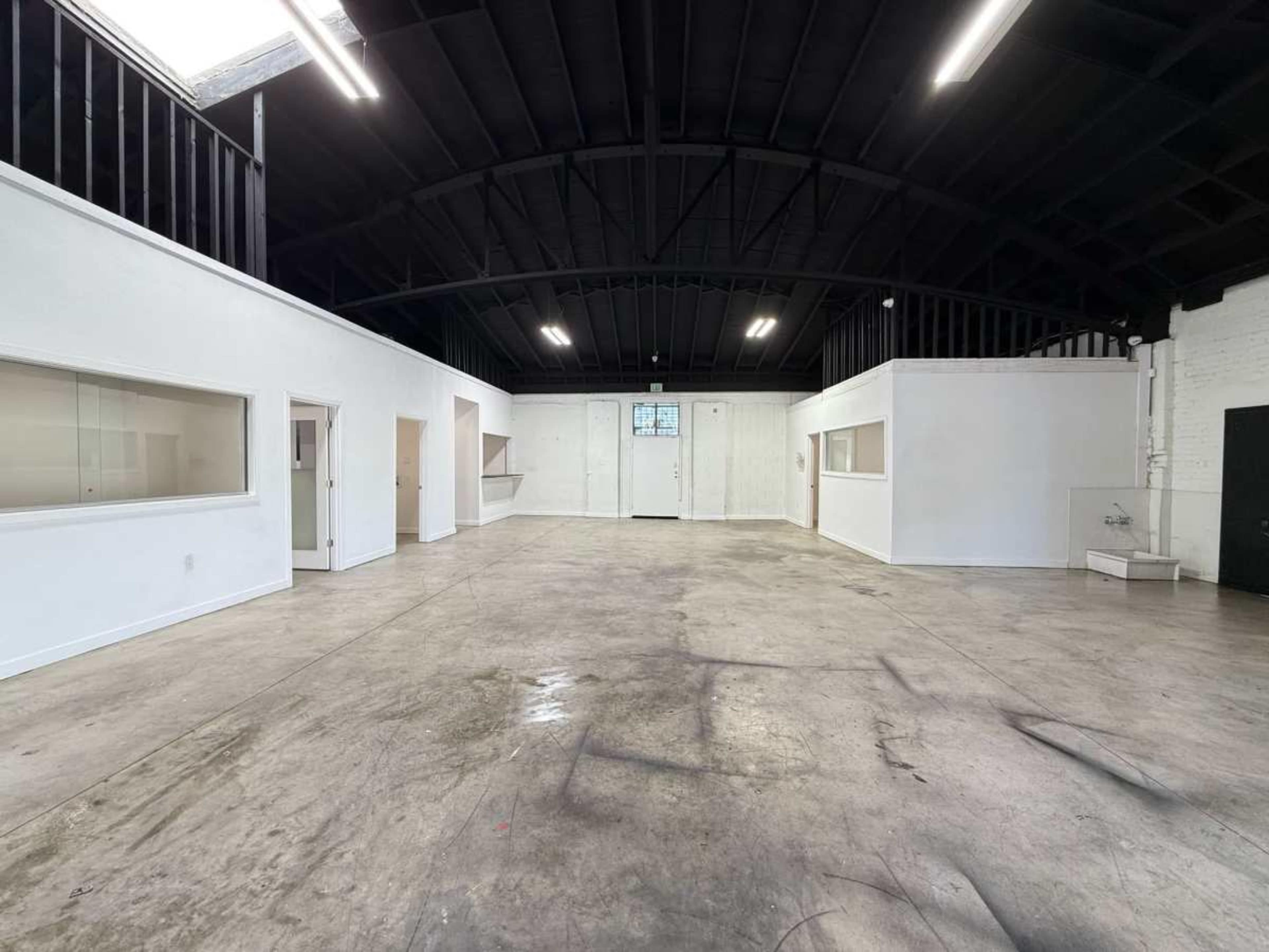 The image shows a spacious, empty room with a high black ceiling, concrete floor, and several white walls and openings.