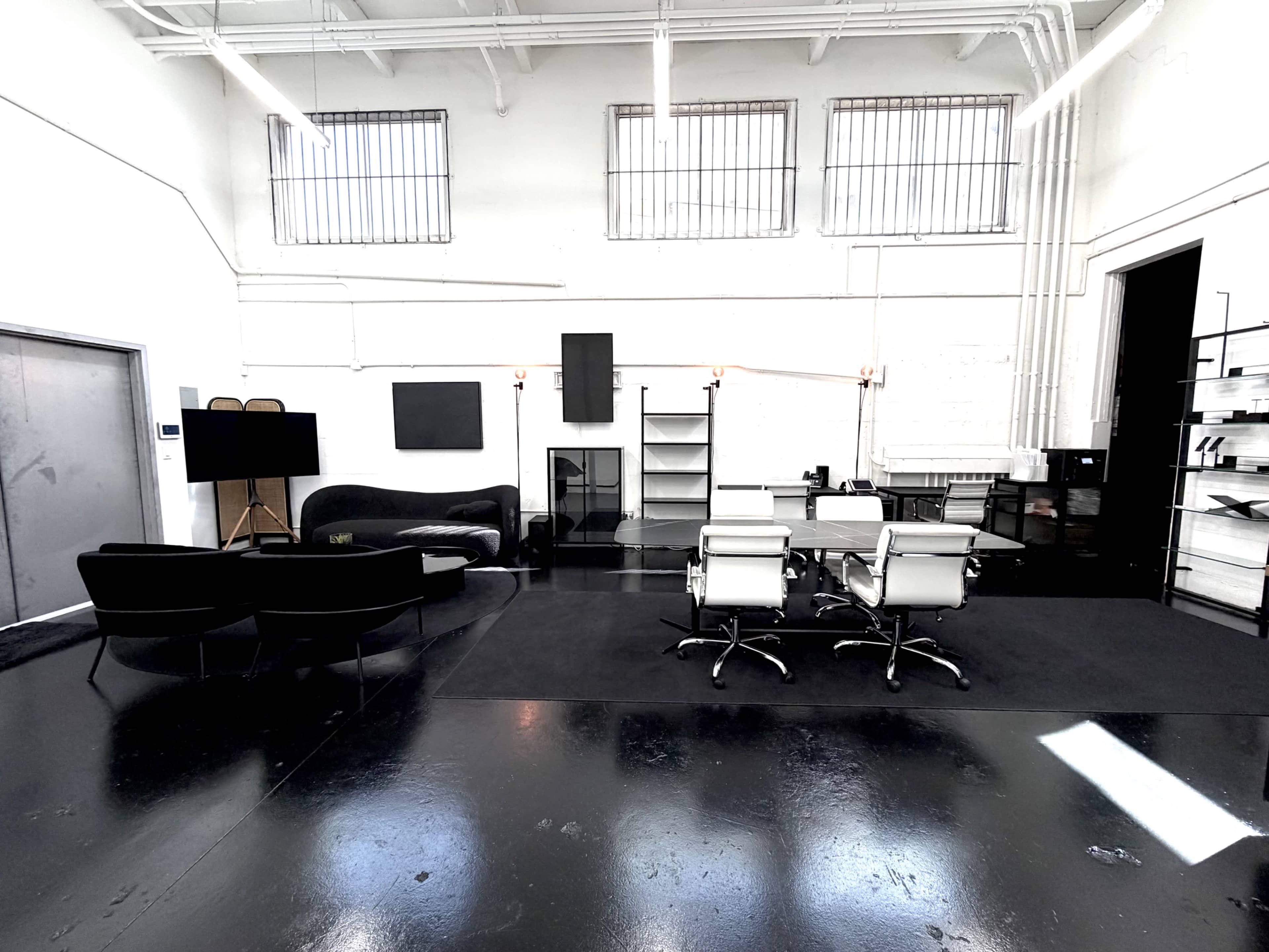 The image shows a spacious, modern office interior featuring a black sofa, two chairs, a conference table with white swivel chairs, and shelves against a white wall.
