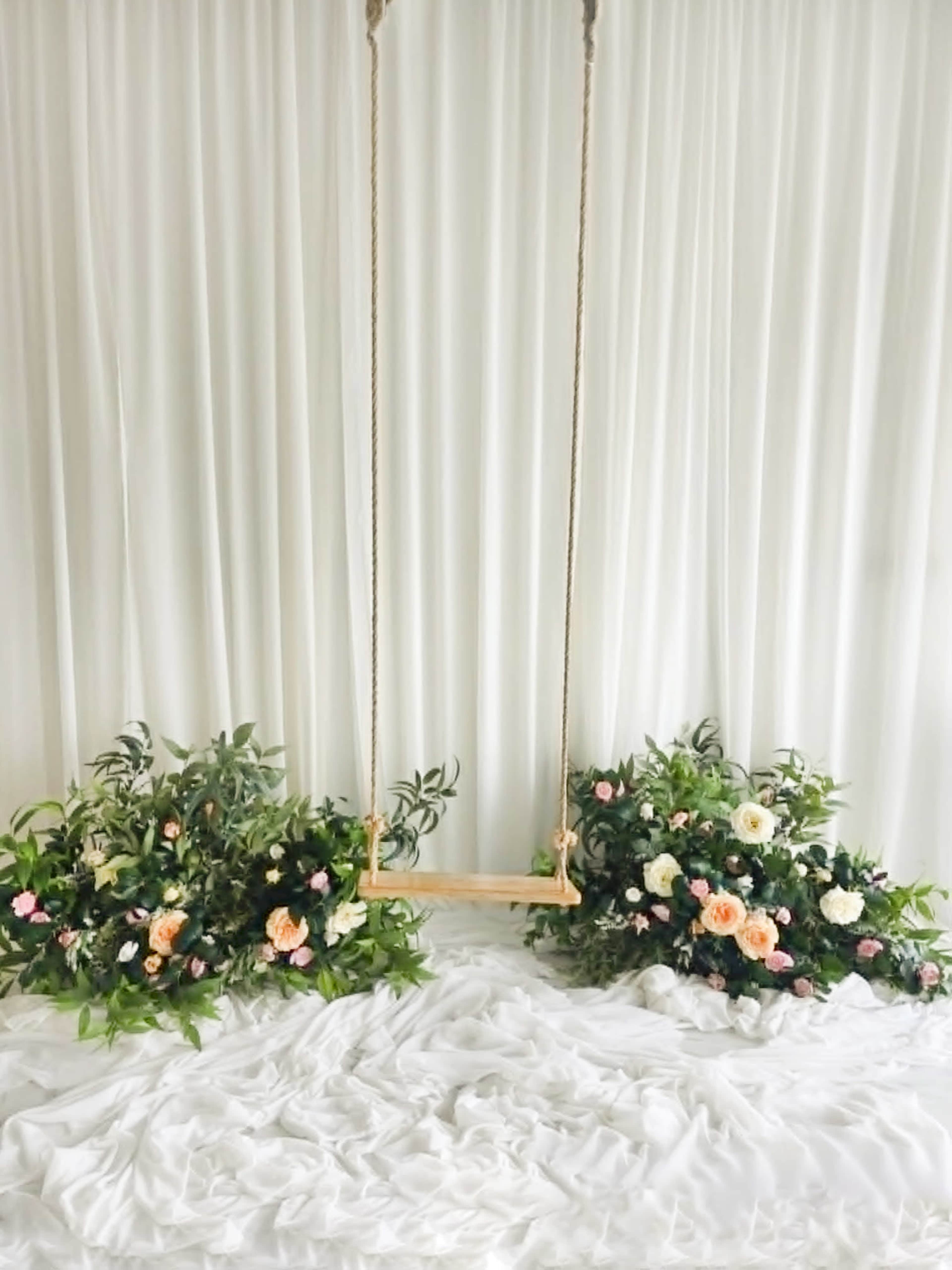 The image shows a wooden swing hanging from a rope surrounded by floral arrangements on both sides, set against a white backdrop.