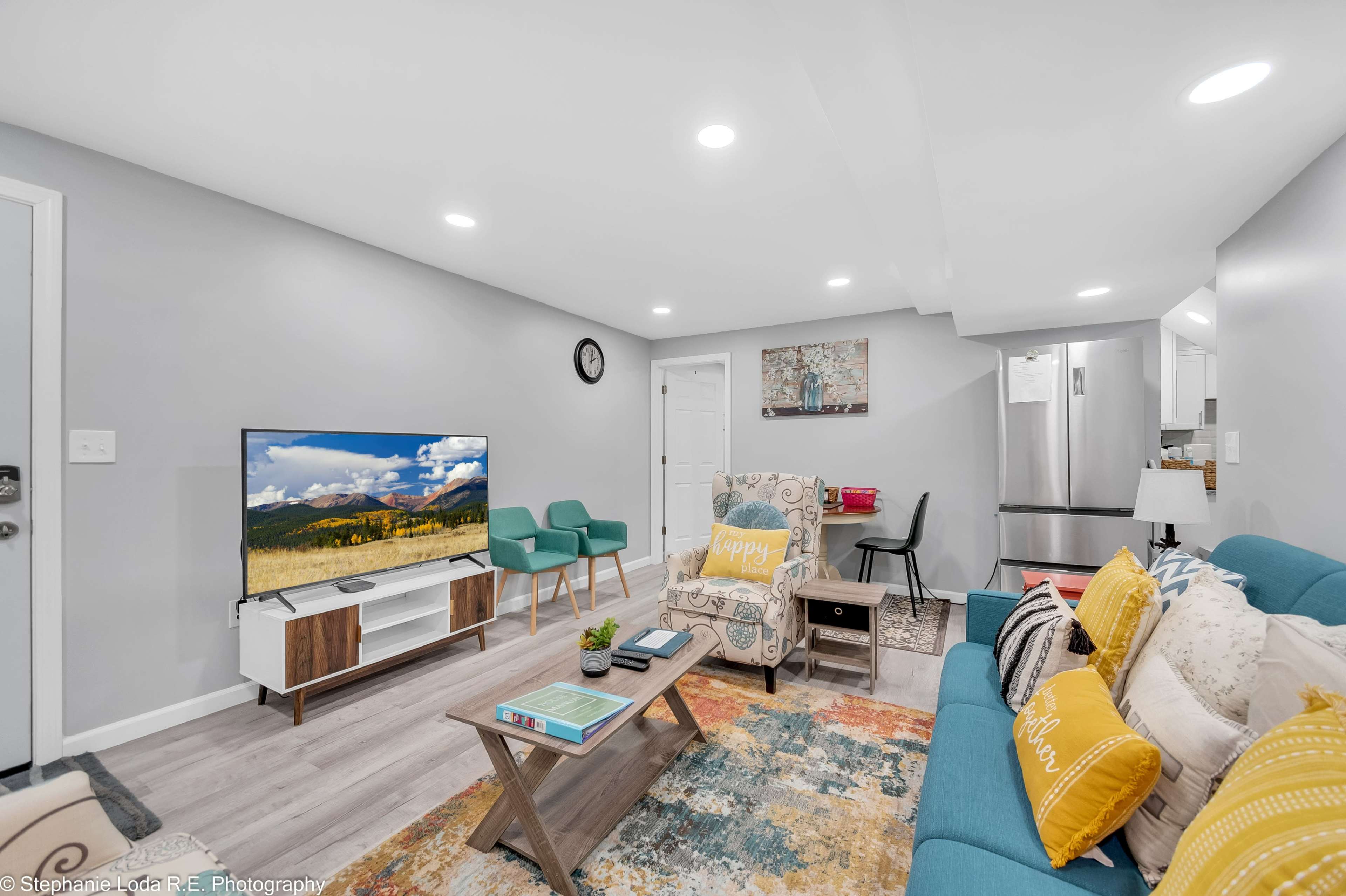 A modern living room featuring a teal sofa, a wooden coffee table, and a television, with various seating and decor elements in a well-lit space.