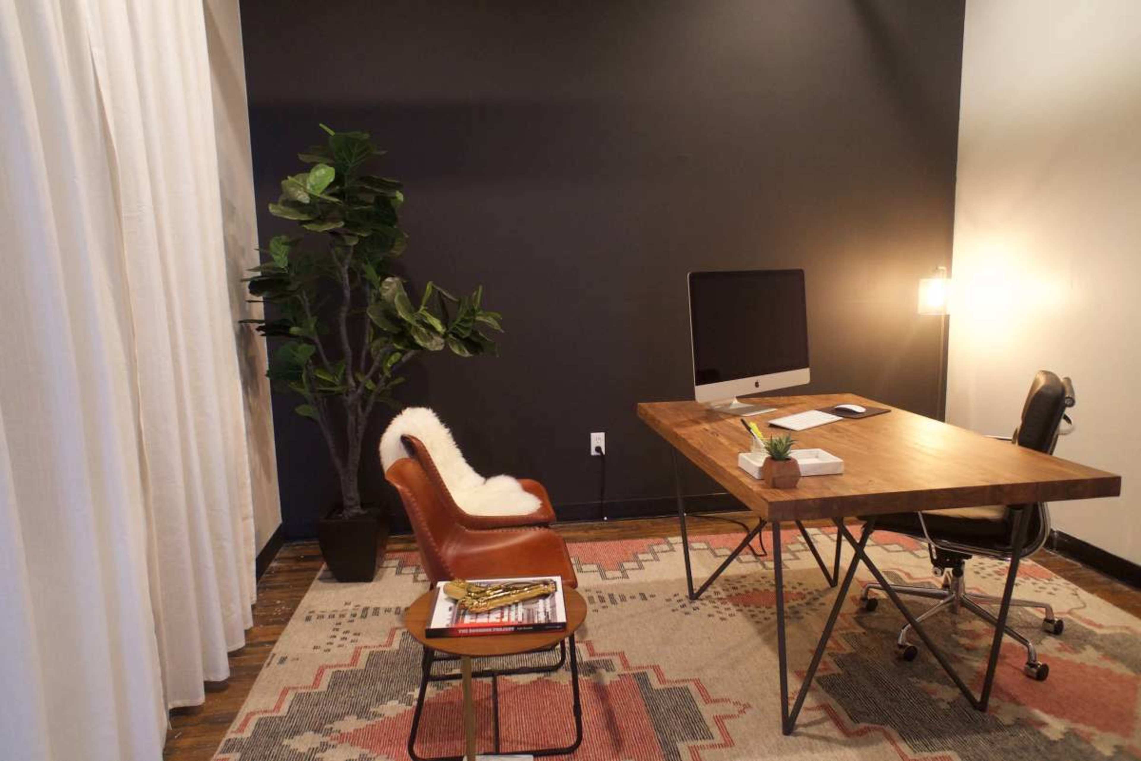 A modern office space features a wooden desk with an iMac, a chair draped with a fur, and a potted plant against a dark wall.