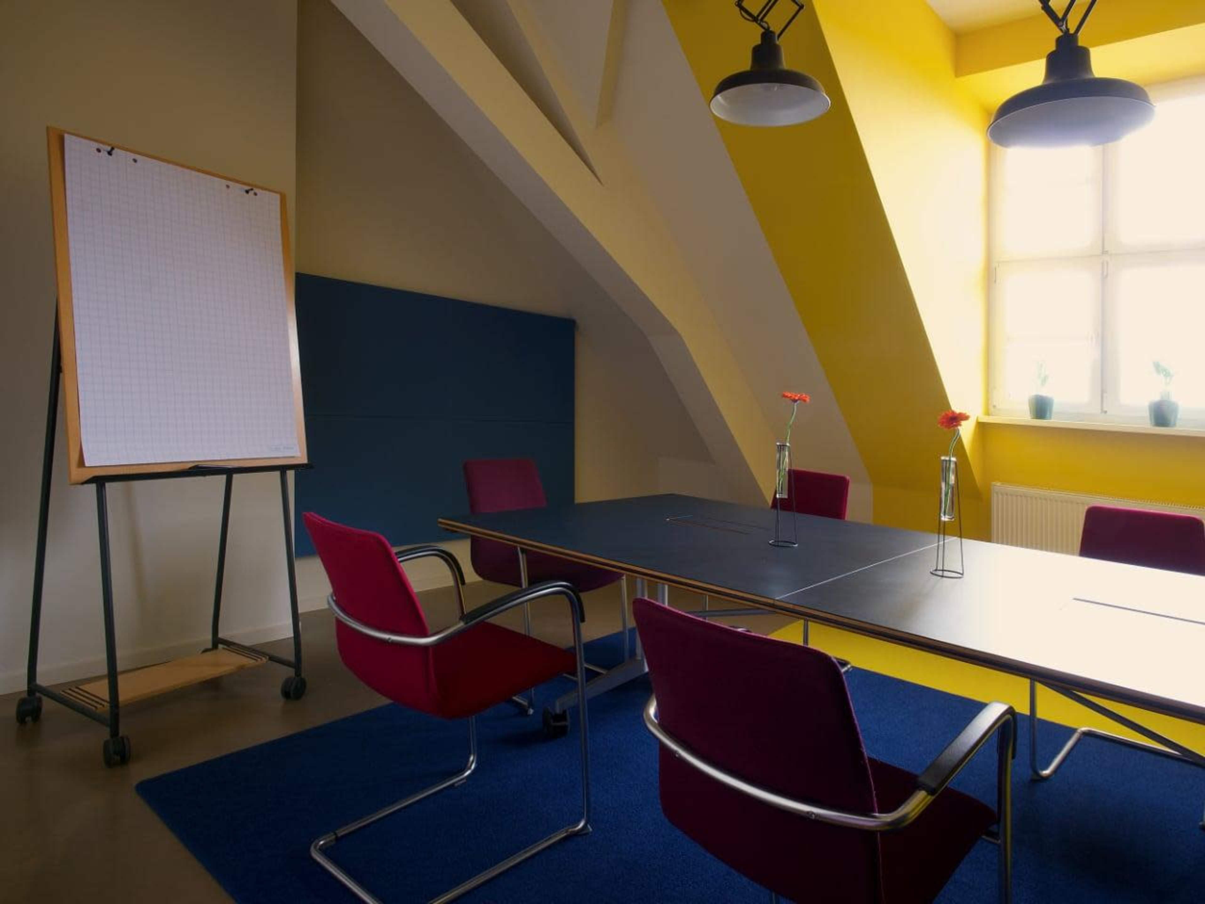 A modern conference room features a long table surrounded by red chairs, a large flip chart on a stand, and bright yellow walls with a window displaying potted plants.