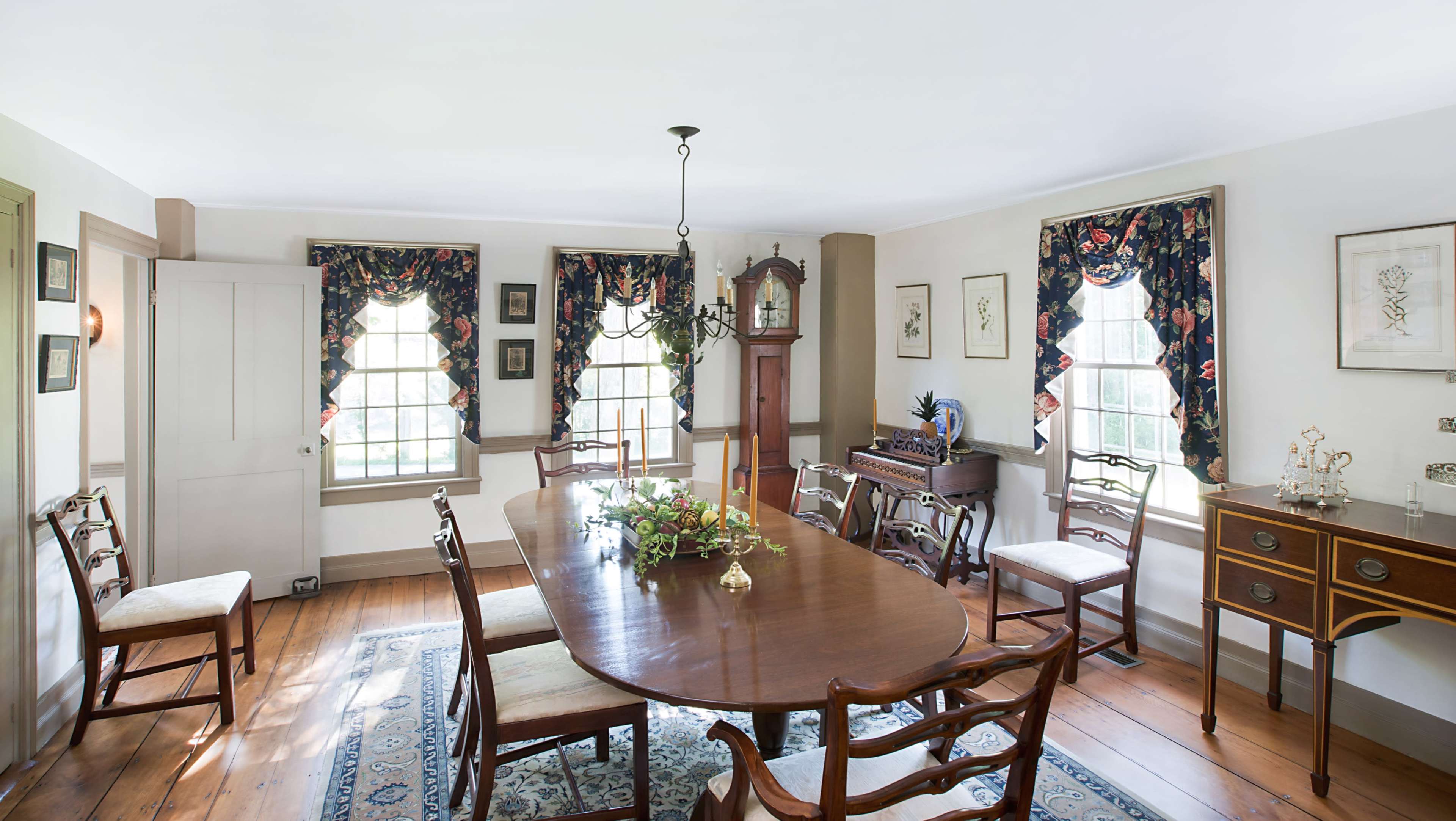The dining room features a long wooden table surrounded by chairs, with decorative curtains and a grandfather clock near the windows.