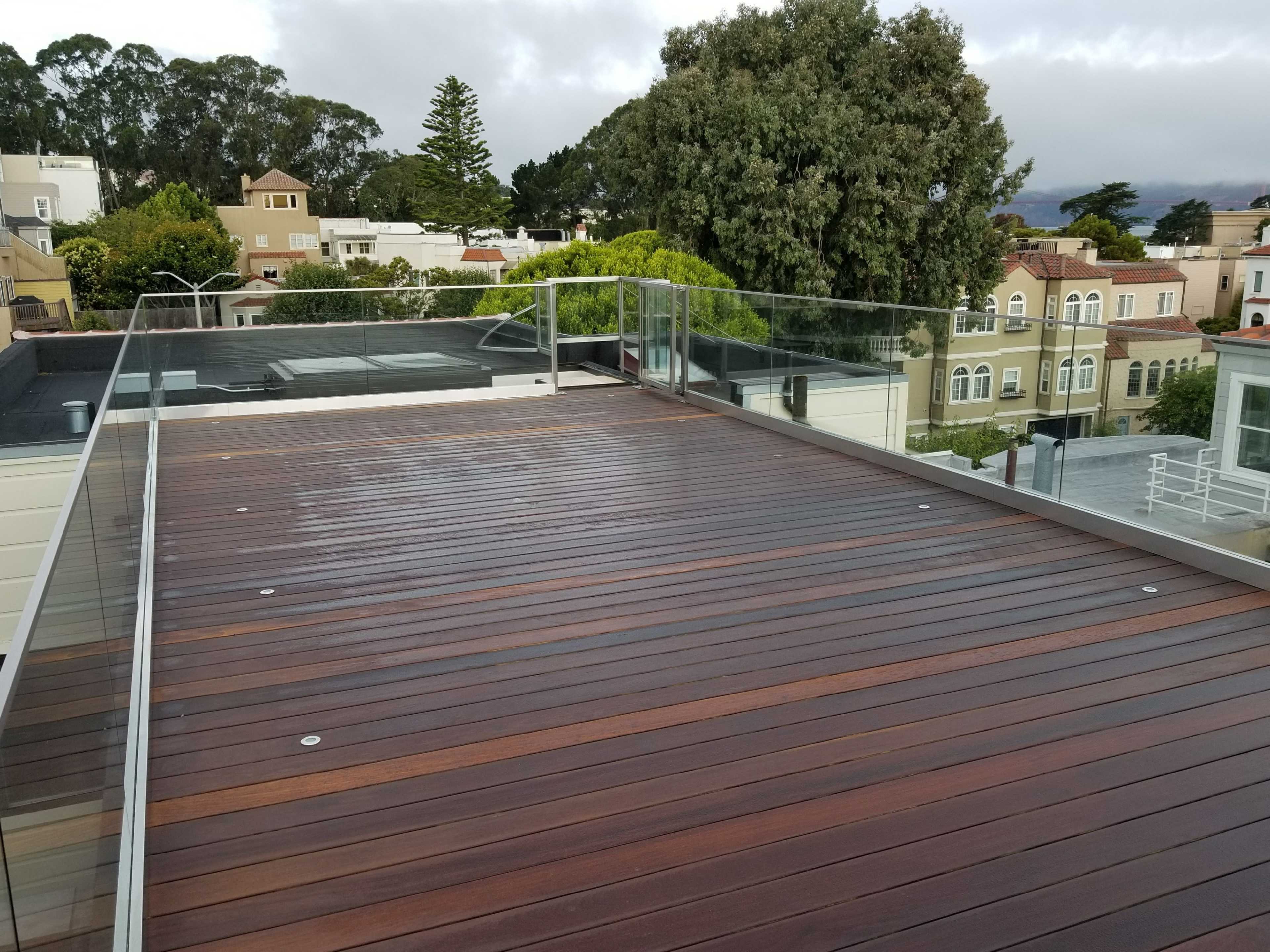 A wooden deck with a glass railing, overlooking a neighborhood of houses and greenery under a cloudy sky.