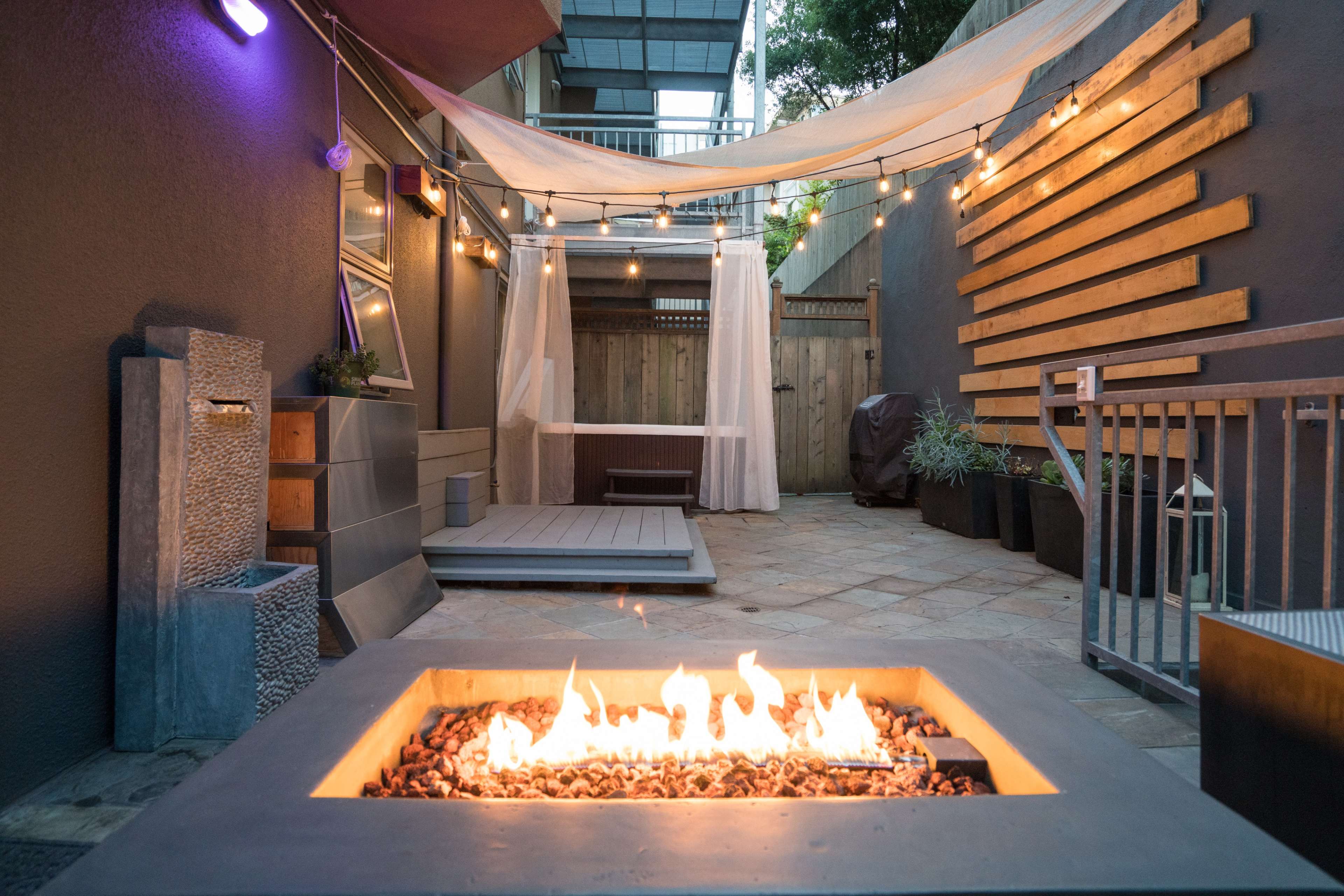 The image shows an outdoor patio area featuring a fire pit surrounded by stone and wooden elements, with string lights overhead and a shaded seating area in the background.