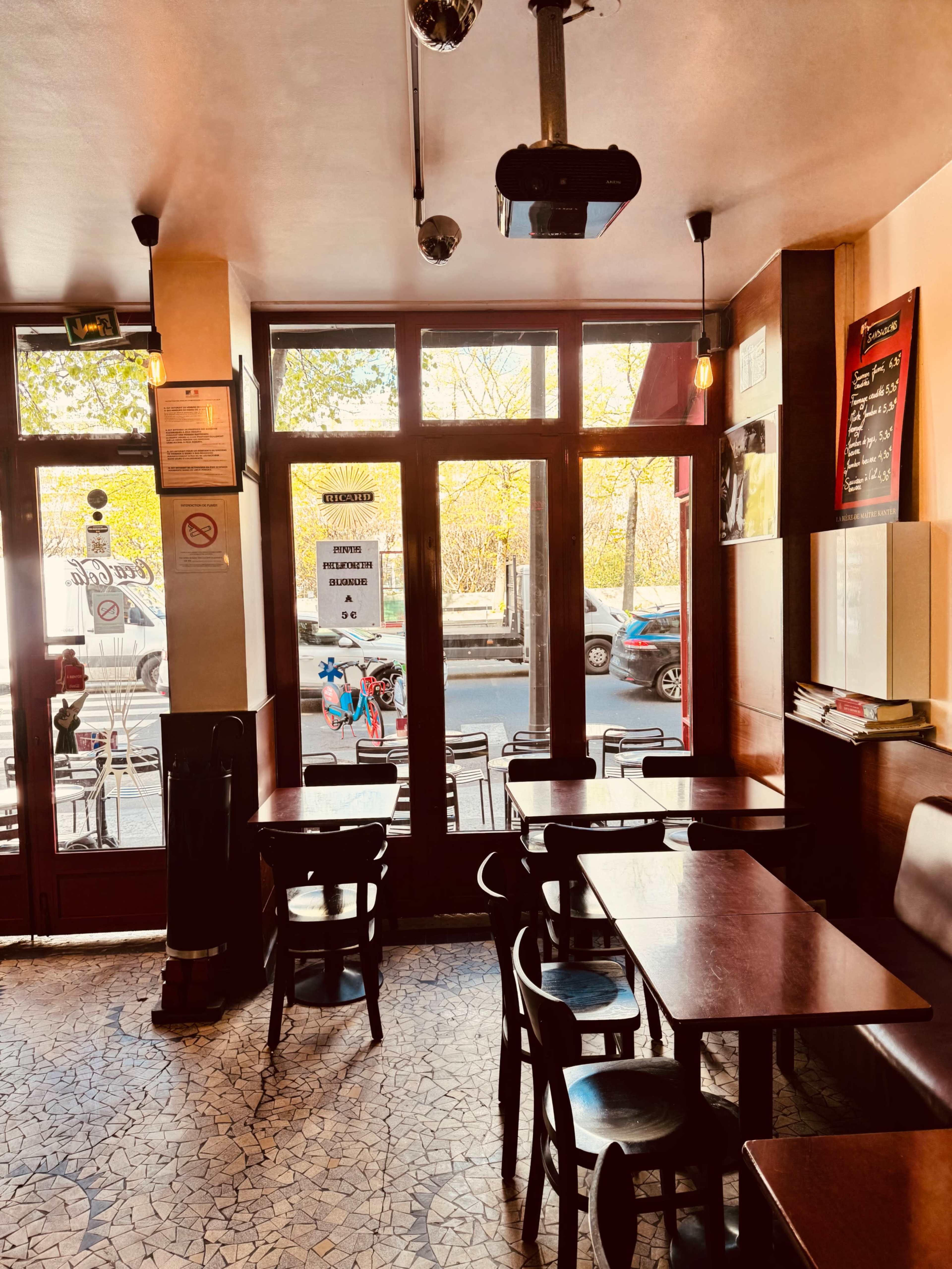 The image shows the interior of a café with wooden tables and chairs, large windows letting in natural light, and a projector mounted on the ceiling.