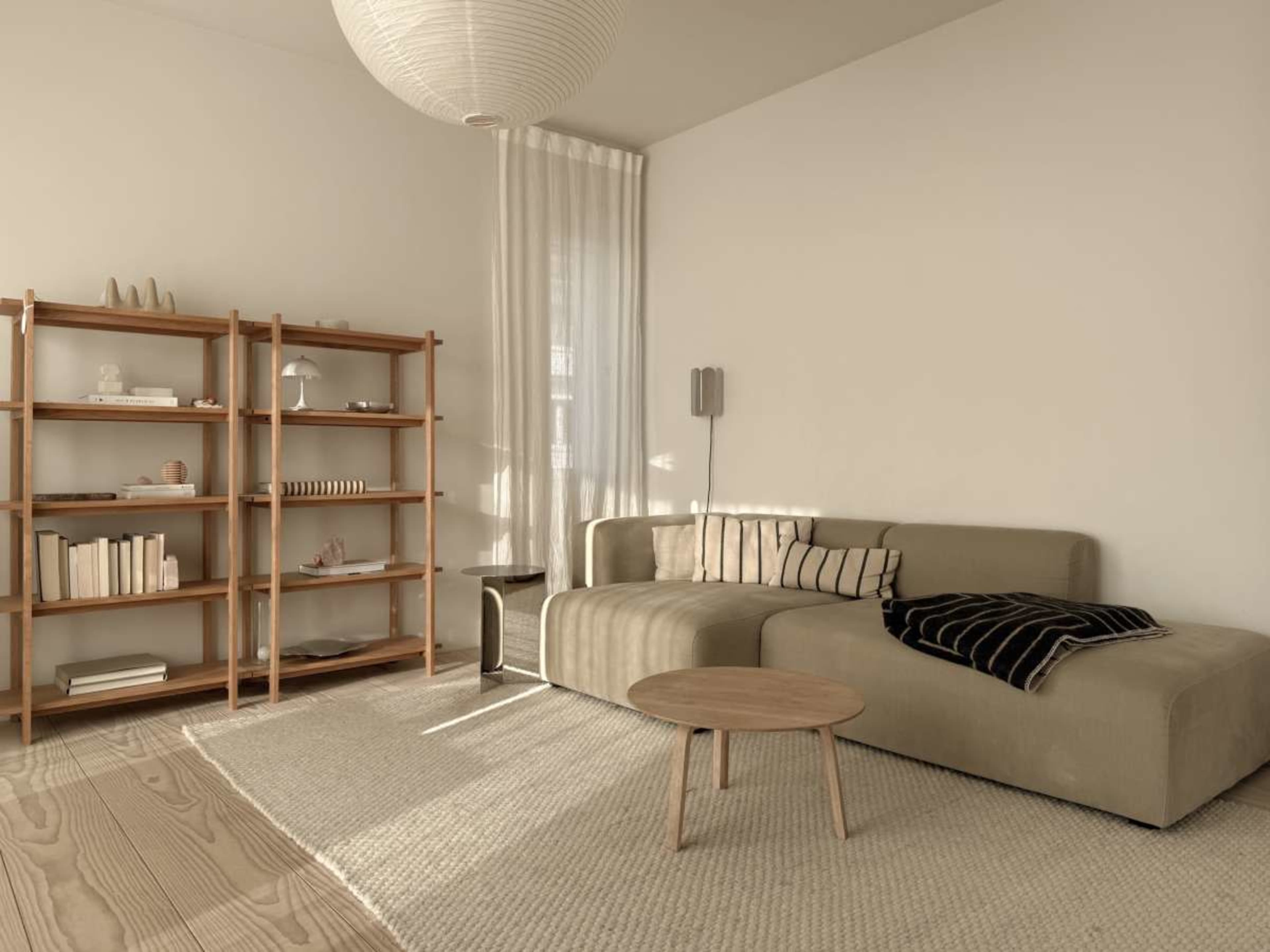 The image shows a minimalist living room with a beige sectional sofa, a wooden bookshelf filled with books and decorative items, and a small round coffee table on a light-colored rug.