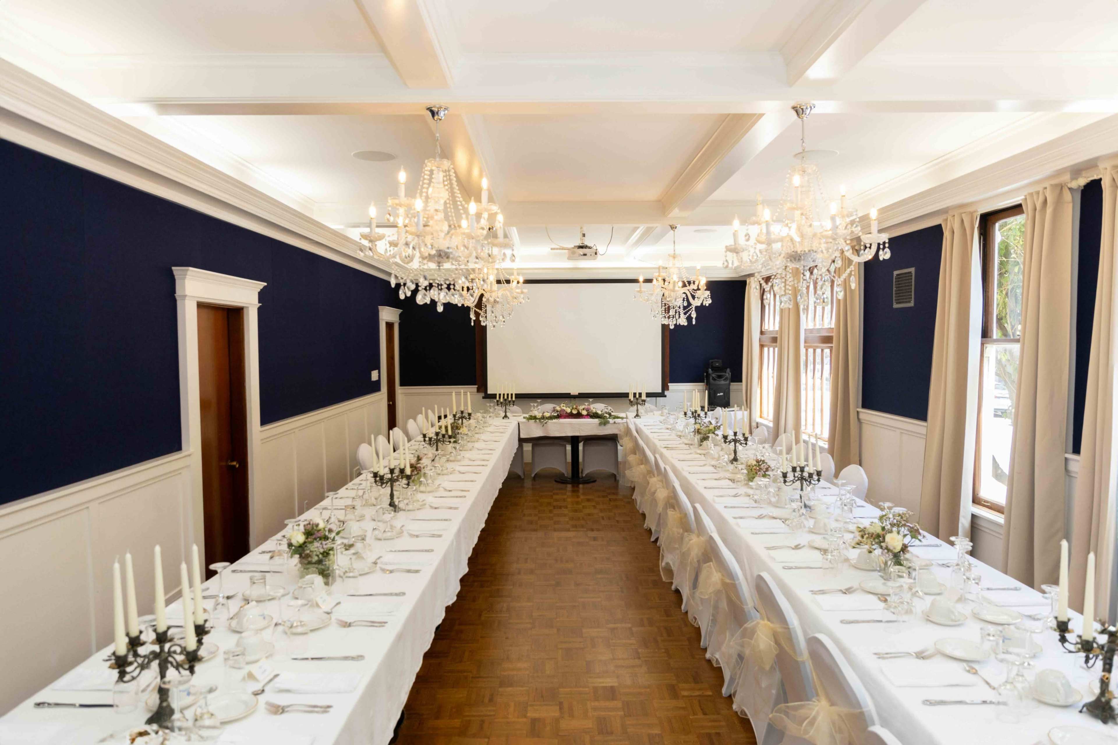 A long banquet table is set with white linens, elegant glassware, and flower arrangements in a well-lit dining room featuring chandeliers and a presentation screen.