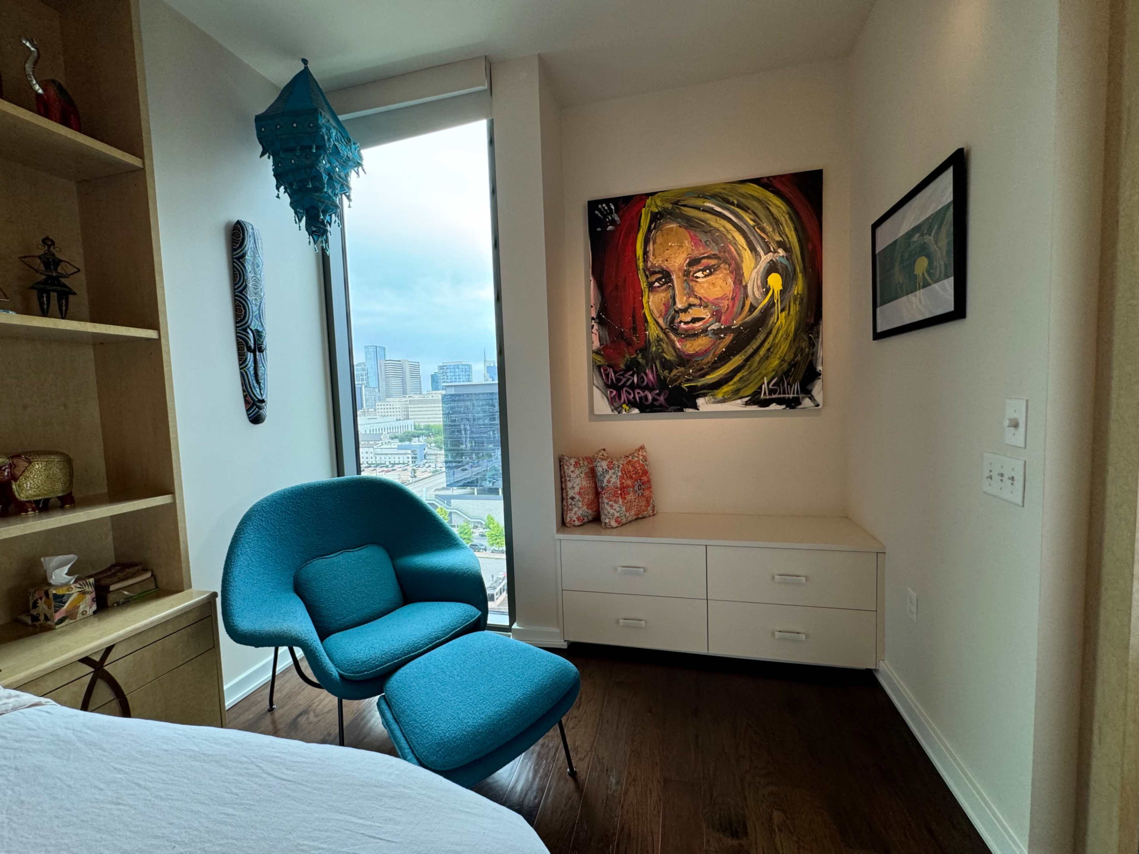 A colorful portrait hangs on the wall above a small white storage unit, beside a blue chair, in a well-lit room with large windows showcasing a city view.