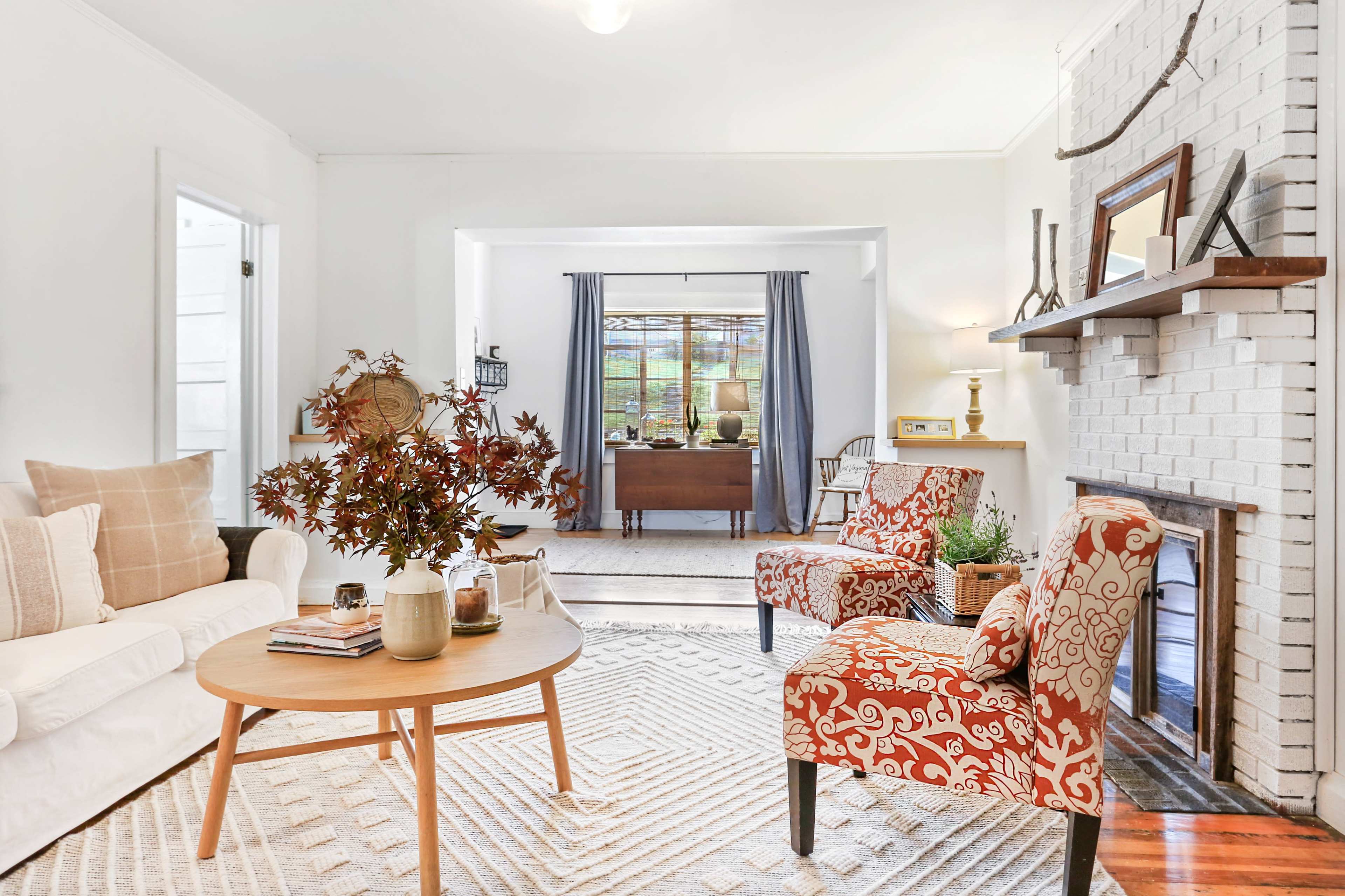 A cozy living room features a white sofa, a round wooden coffee table, and two chairs with patterned upholstery, with a view of a workspace through an open archway.