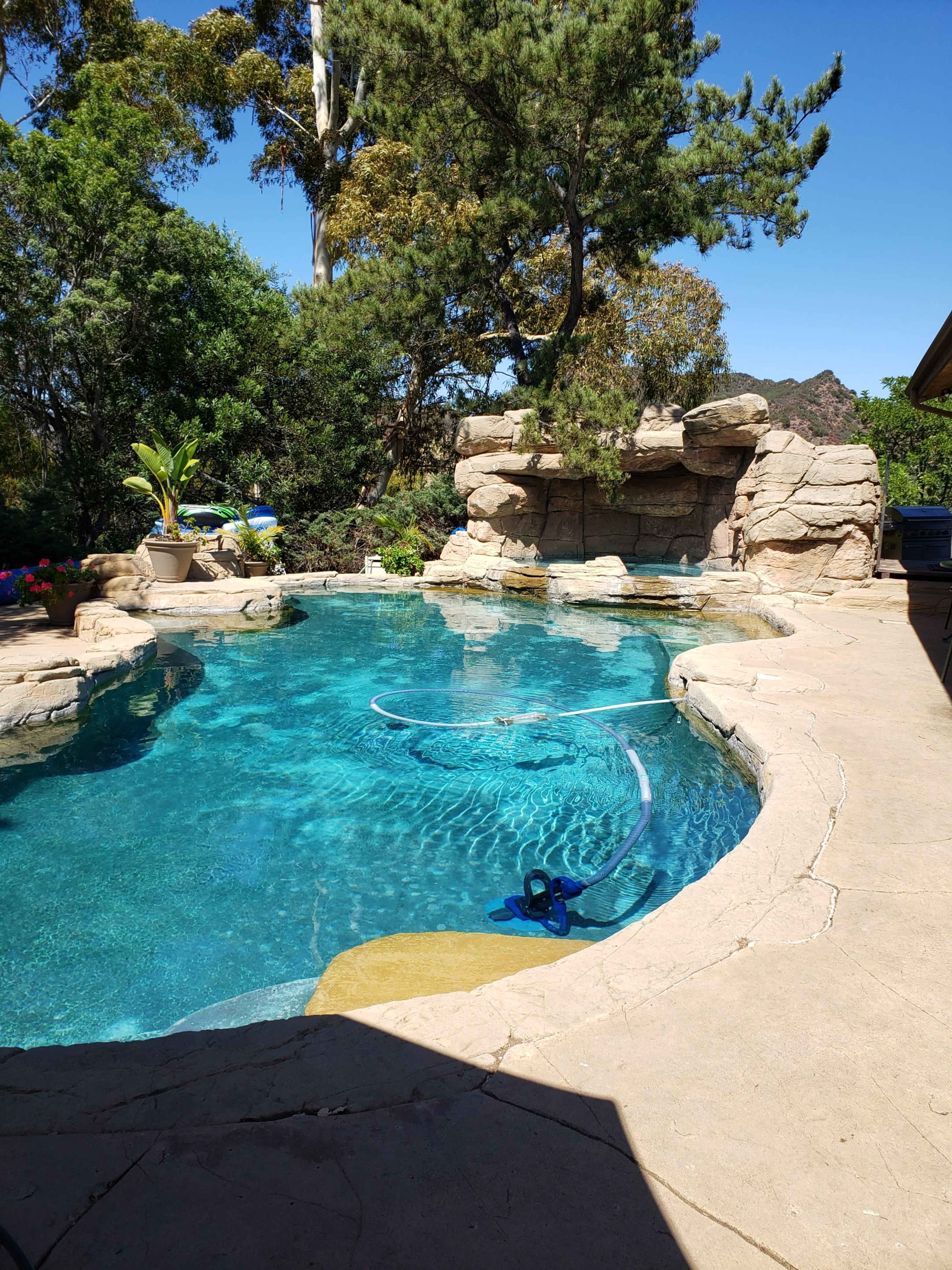 The image shows a backyard swimming pool surrounded by rocks and greenery, with a pool cleaner visible in the water.