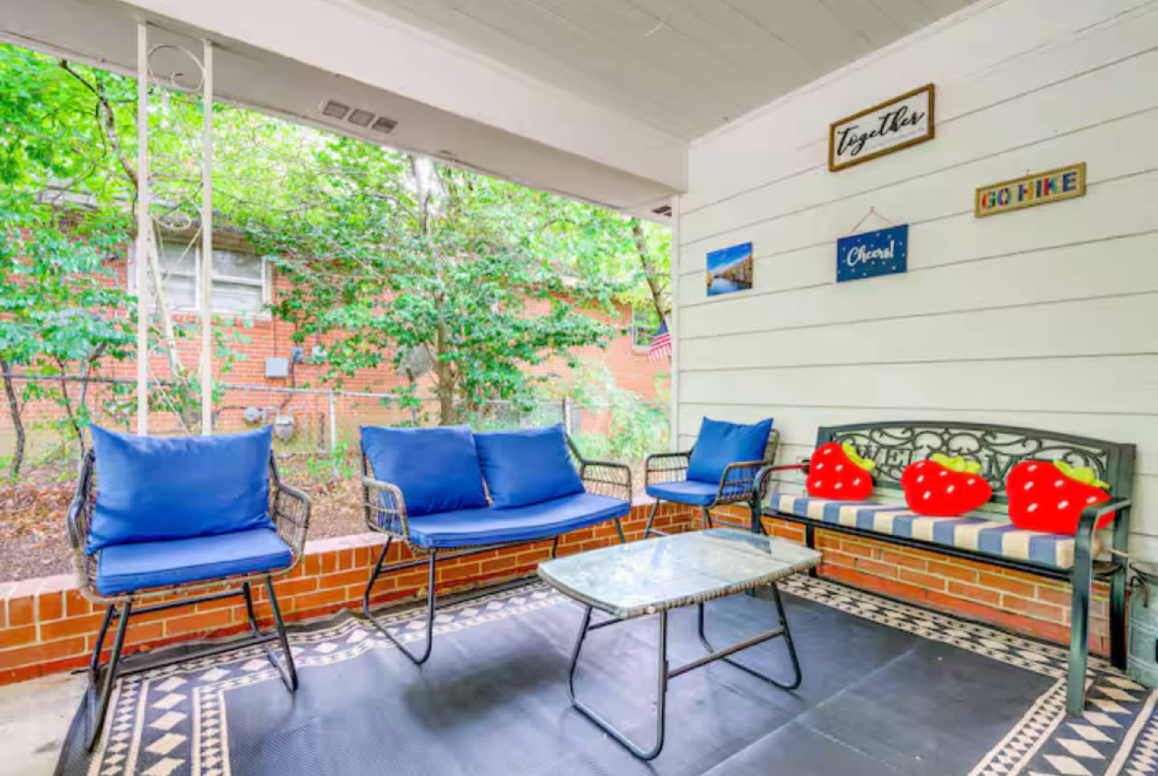 The image shows a covered porch with blue seating, a glass coffee table, and decorative signs against a backdrop of greenery.