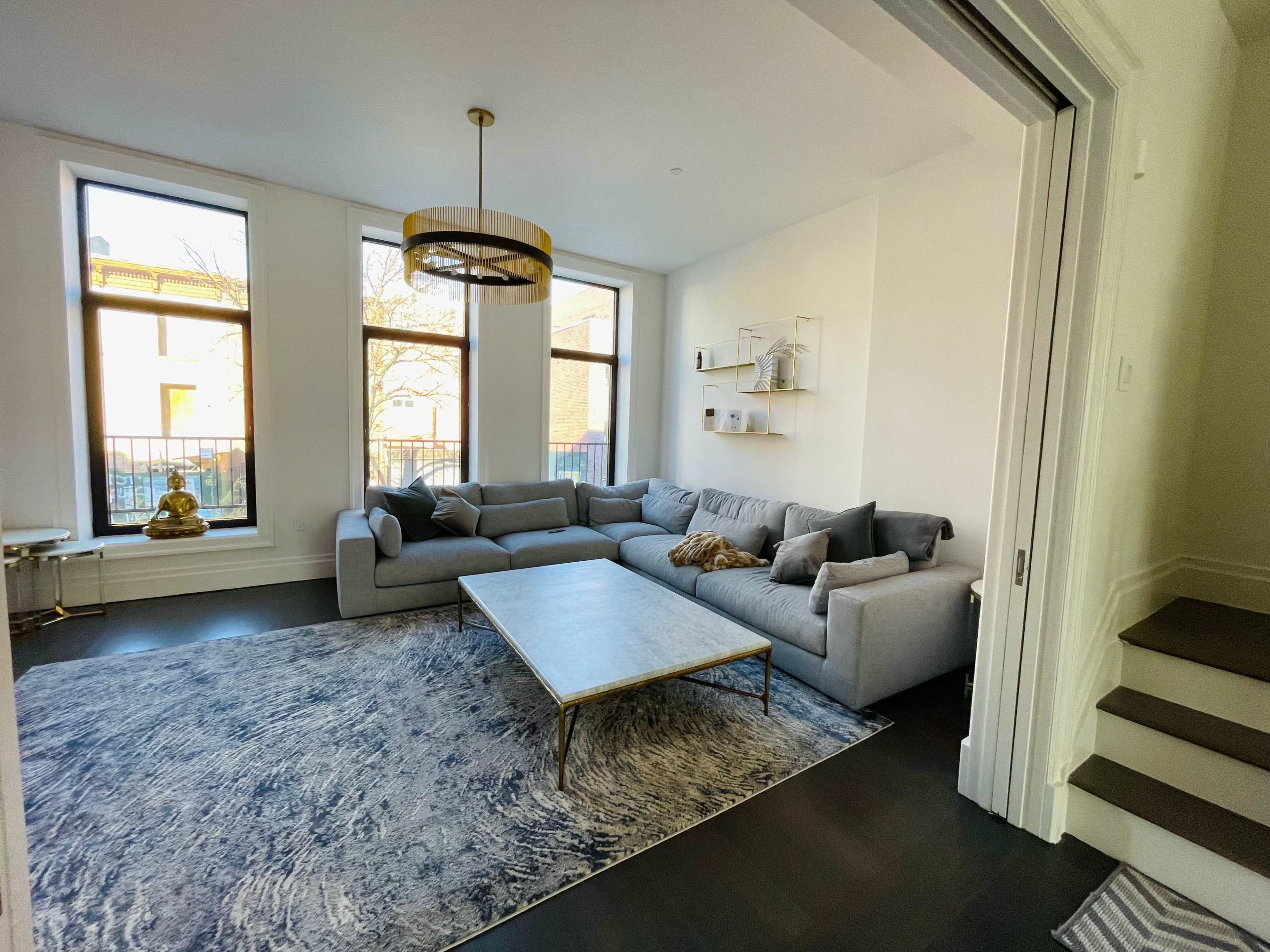 A modern living room features a large gray sectional sofa, a rectangular coffee table, and natural light streaming through three tall windows.