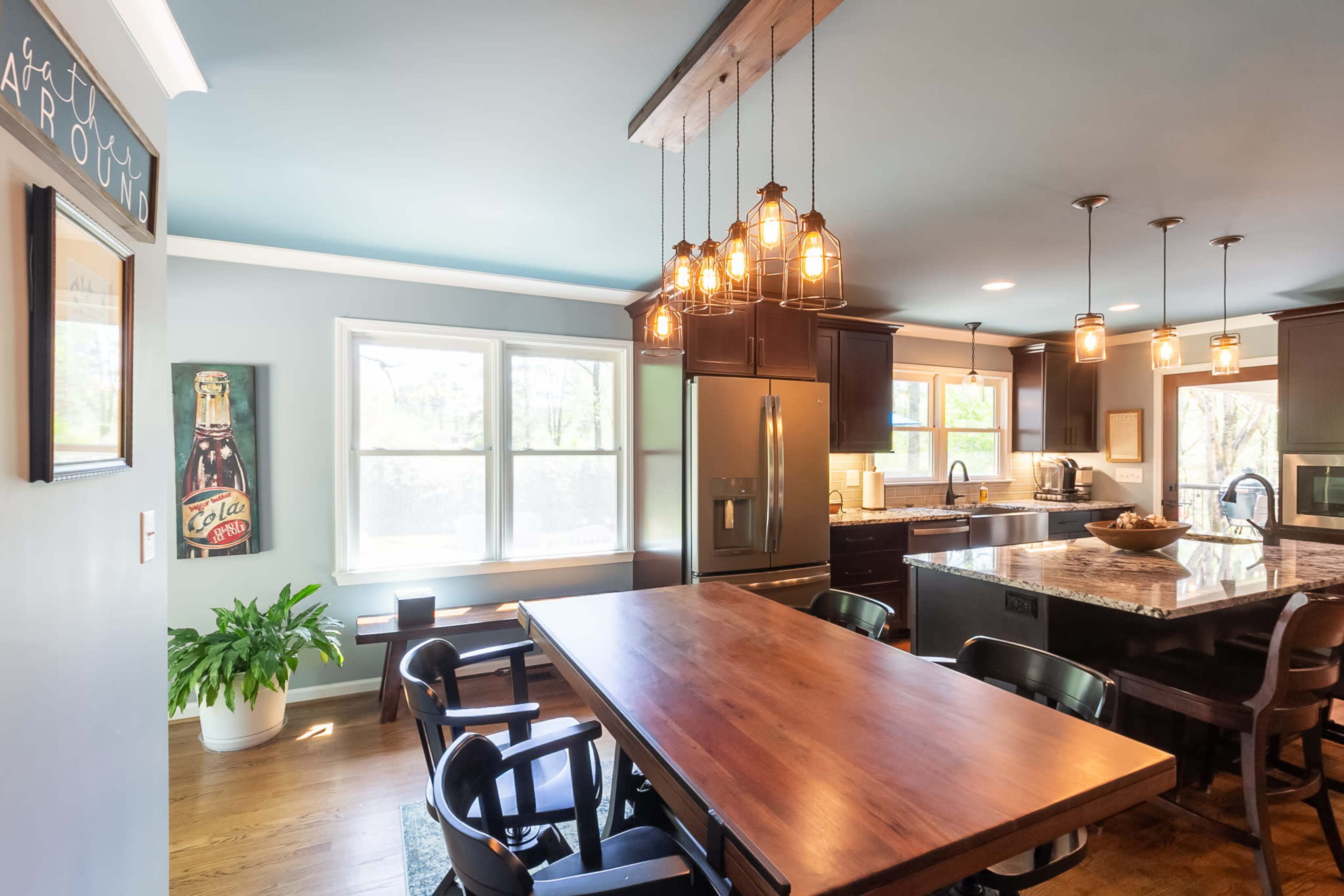 A modern kitchen with a wooden dining table, pendant lights, and dark cabinetry, featuring a large window that allows natural light to enter.