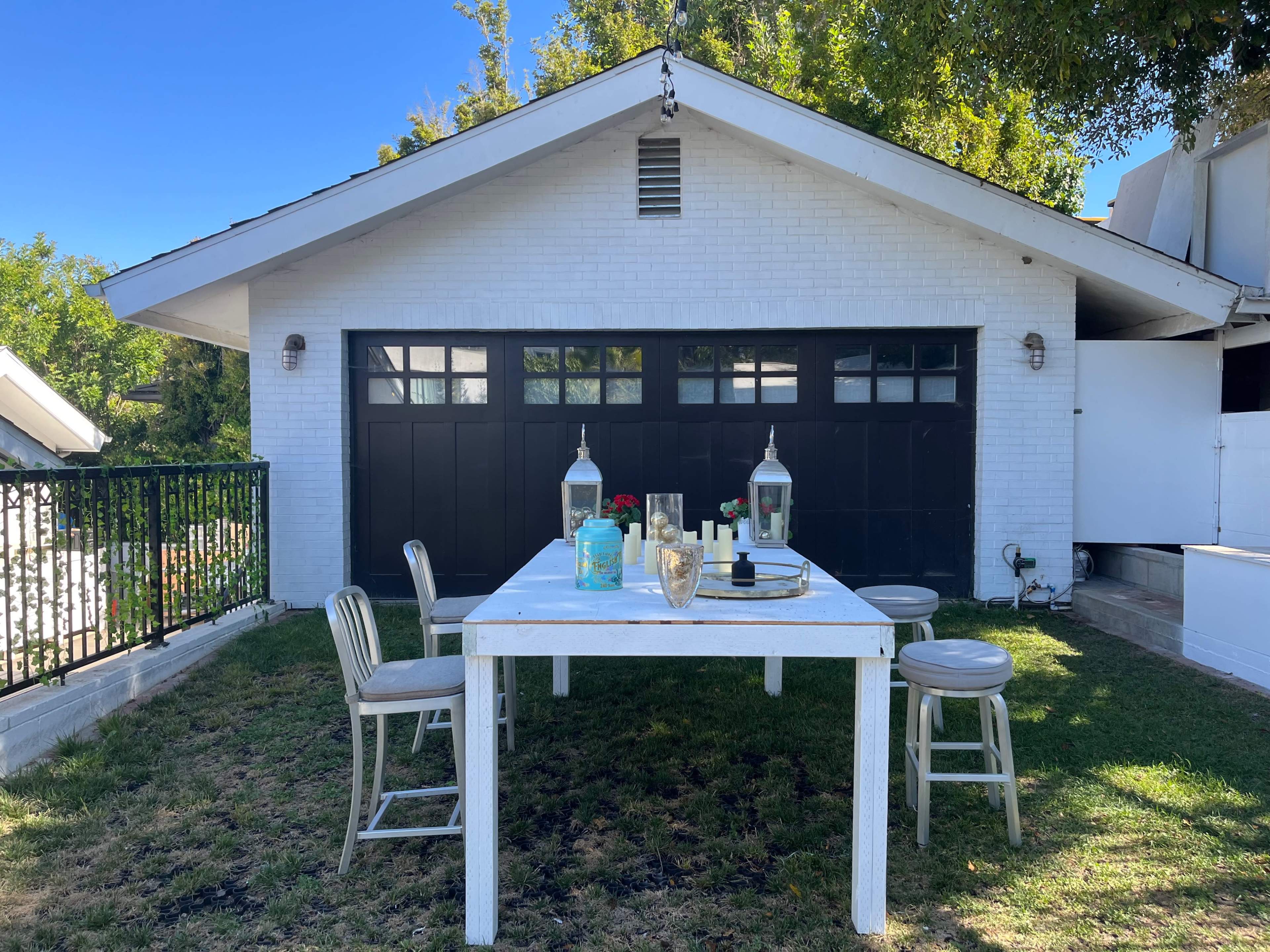 A white table with seating is placed on grass next to a garage with a black door, surrounded by greenery.