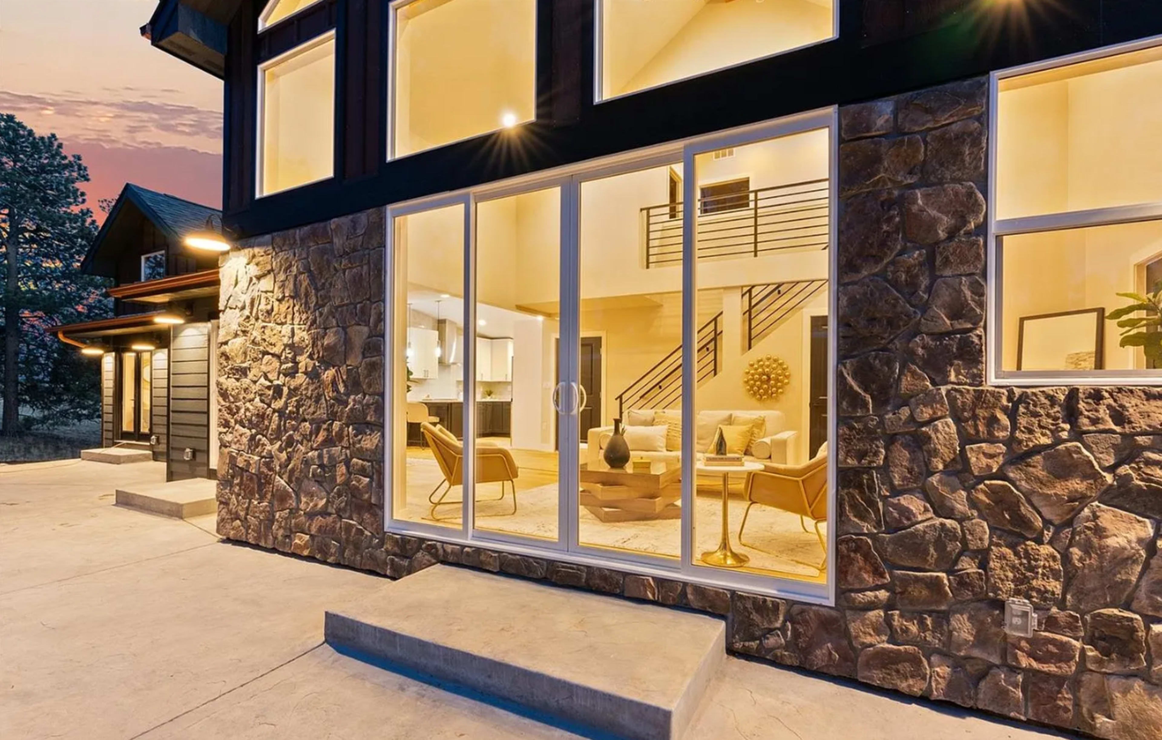 The image shows a modern stone house with large glass windows revealing a warmly lit living space and an outdoor area at dusk.