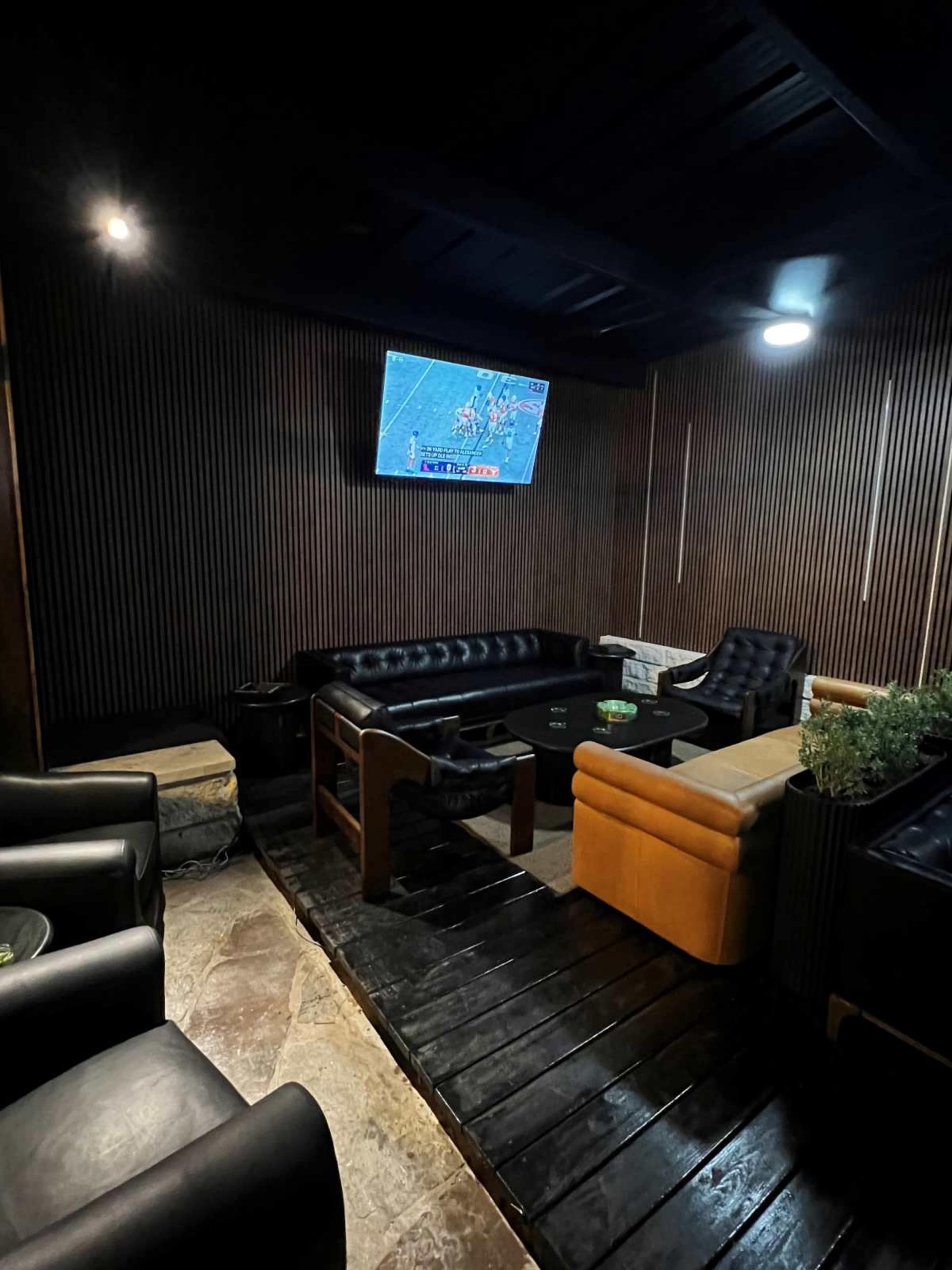 The image shows a dimly lit lounge area with leather couches, a wall-mounted TV, and wood paneling.