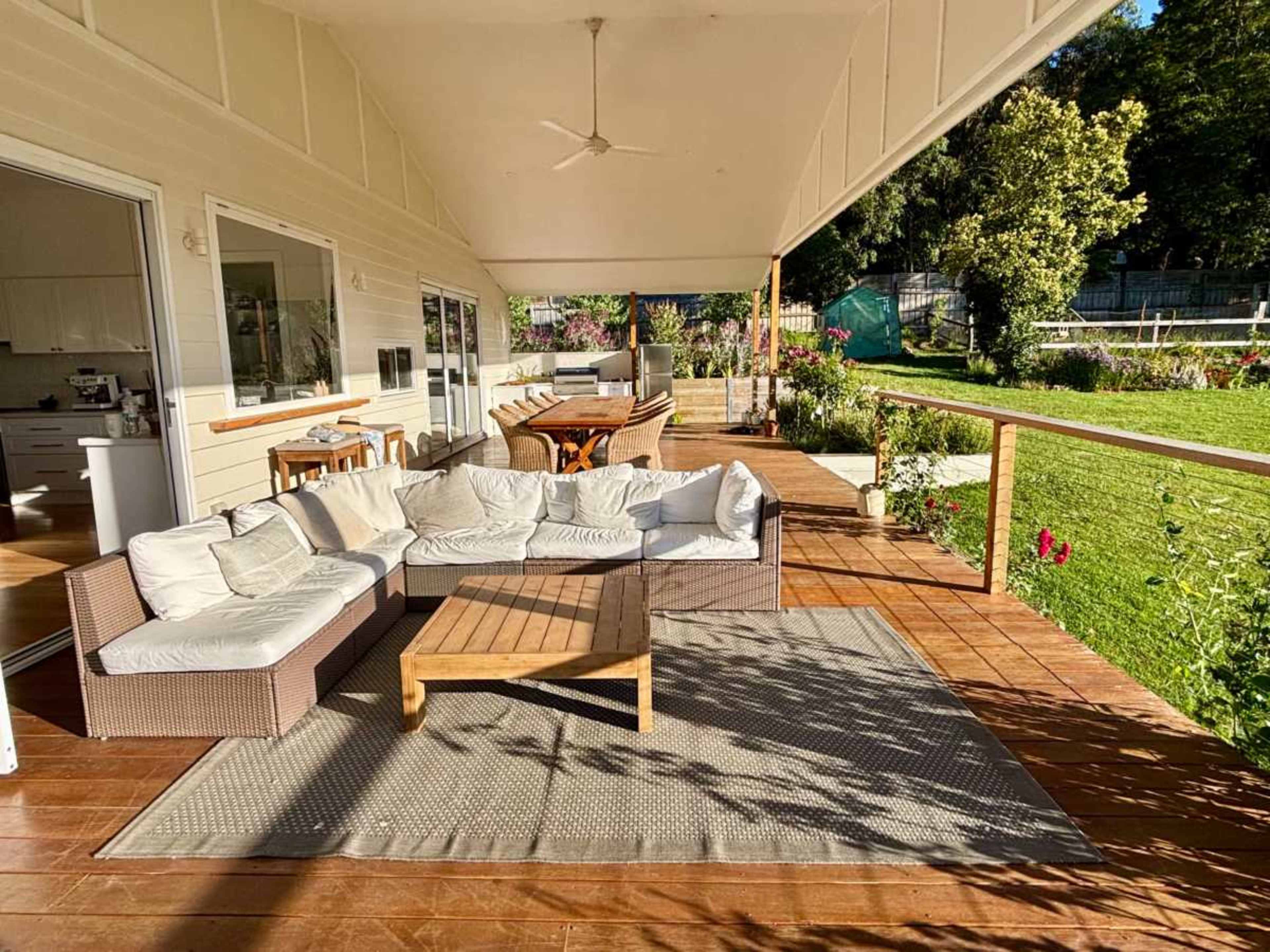 The image shows a spacious outdoor porch with a comfortable sectional sofa, a wooden coffee table, and greenery in the background.