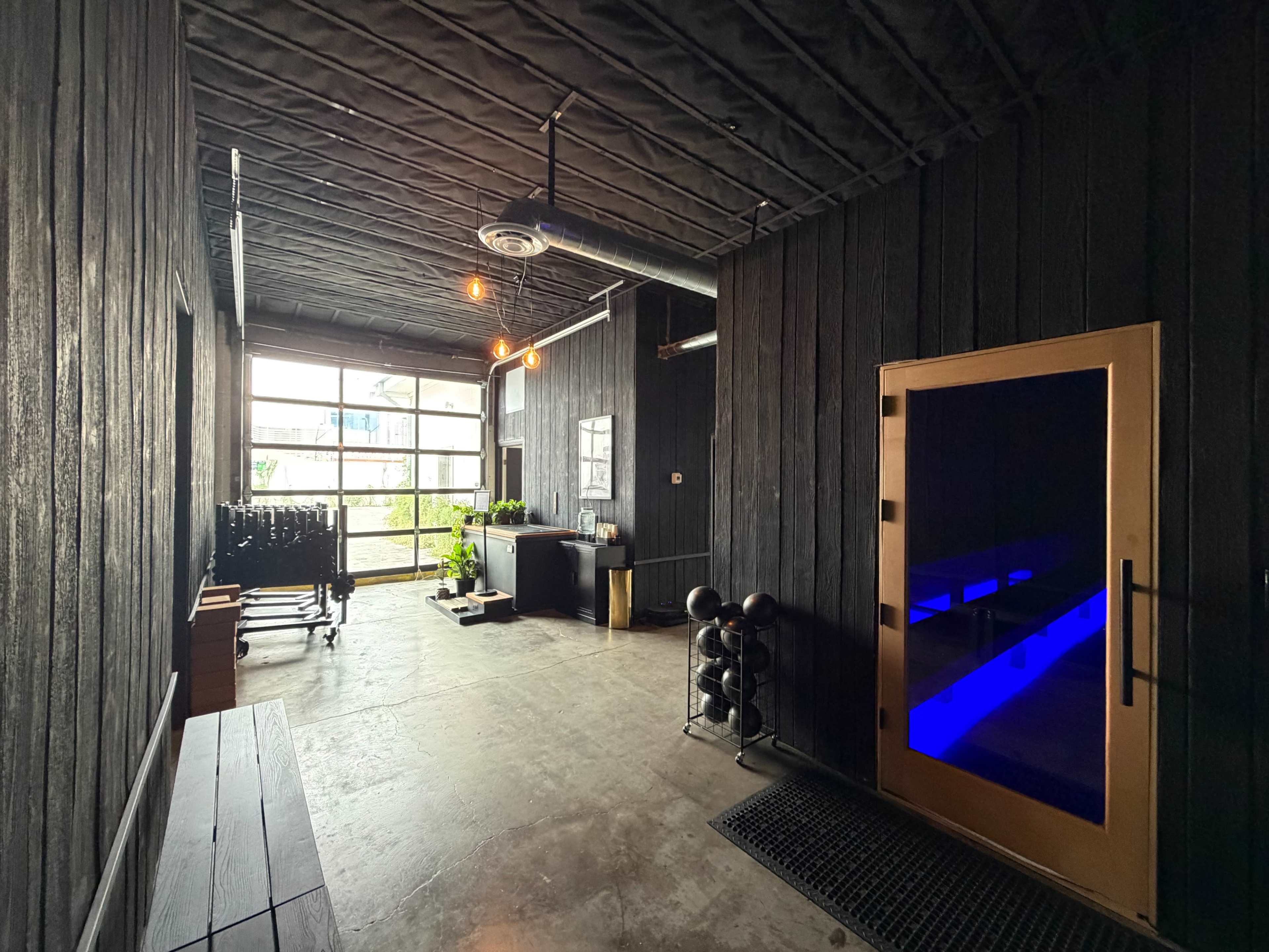 The image shows a modern hallway with dark wooden walls leading to a large window, a small reception area, and a sauna with blue lighting.