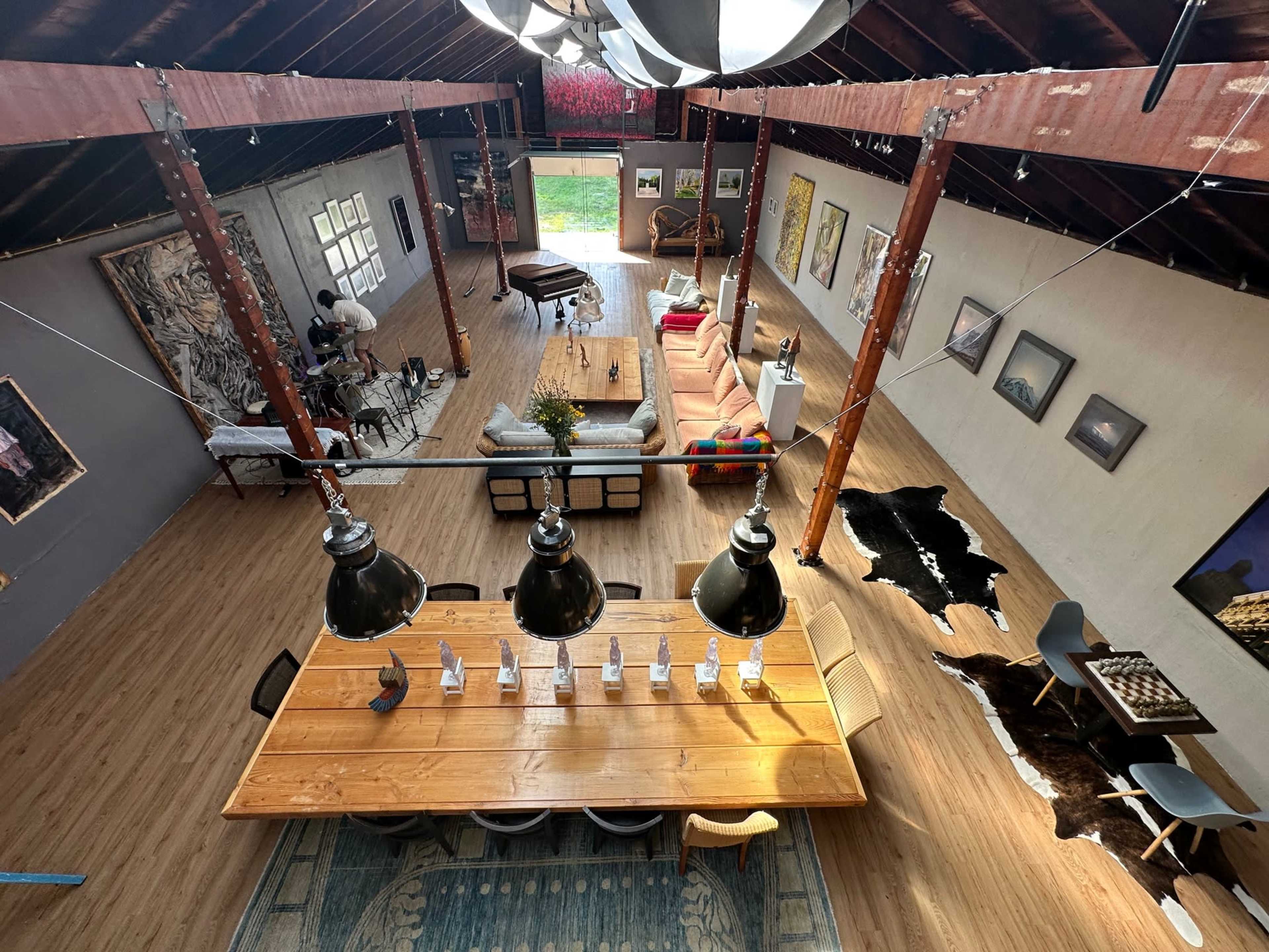 A spacious interior features a long wooden dining table under pendant lights, surrounded by artwork, seating areas, and large windows.