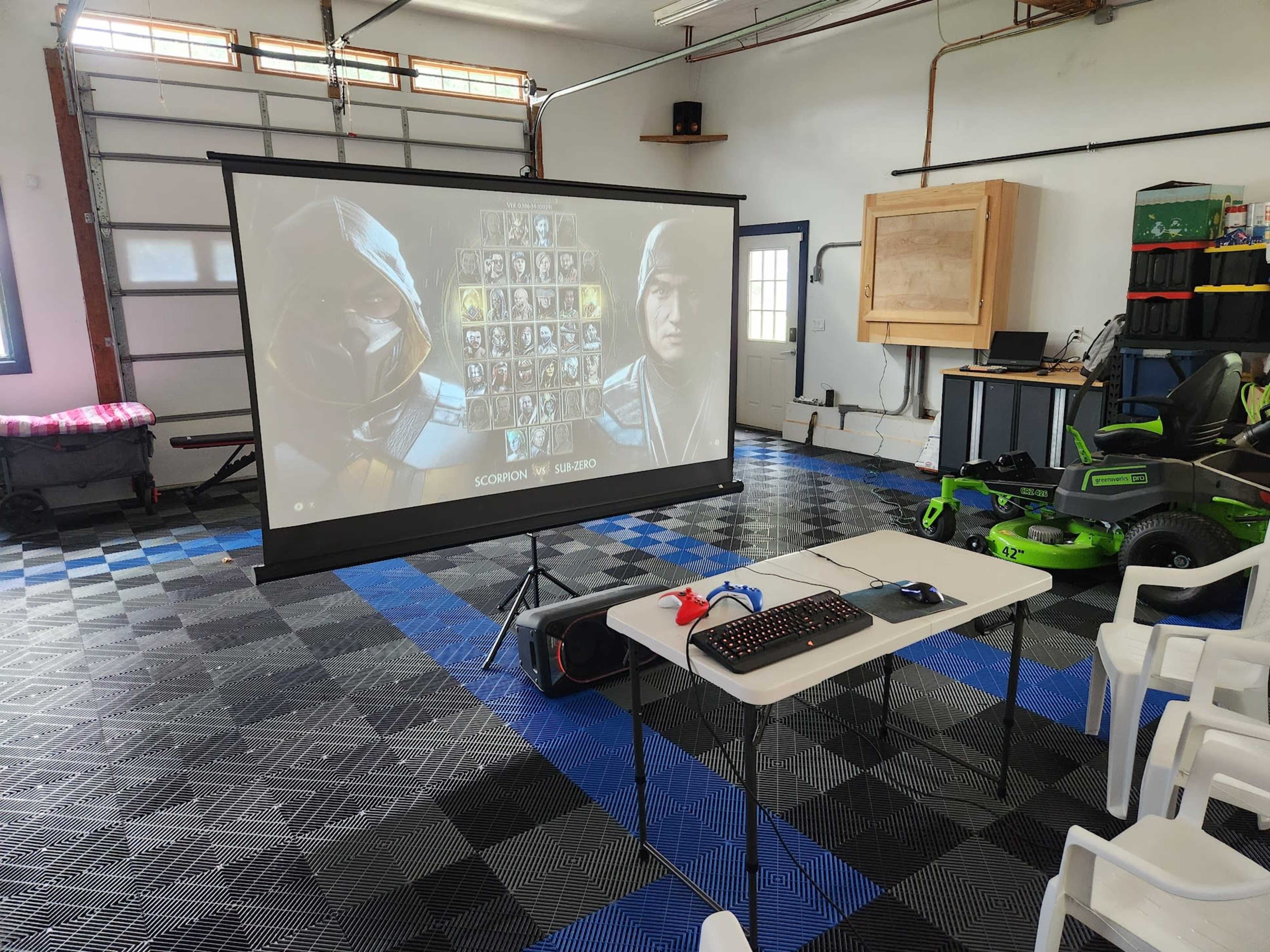 A home garage is set up with a large projection screen displaying a video game character selection menu, a table with a keyboard and gaming controls, and chairs nearby.