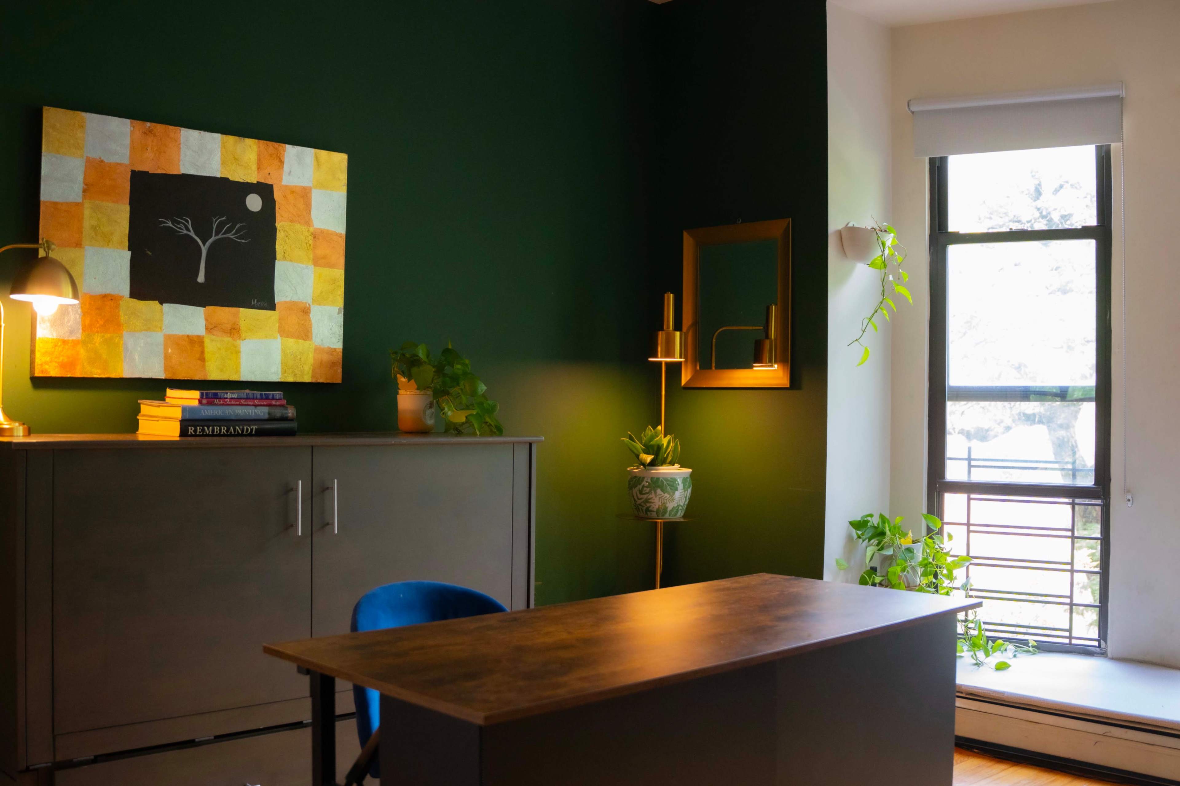 The image depicts a modern office space with a dark green wall, a wooden desk, a colorful wall painting, and plants near a window.