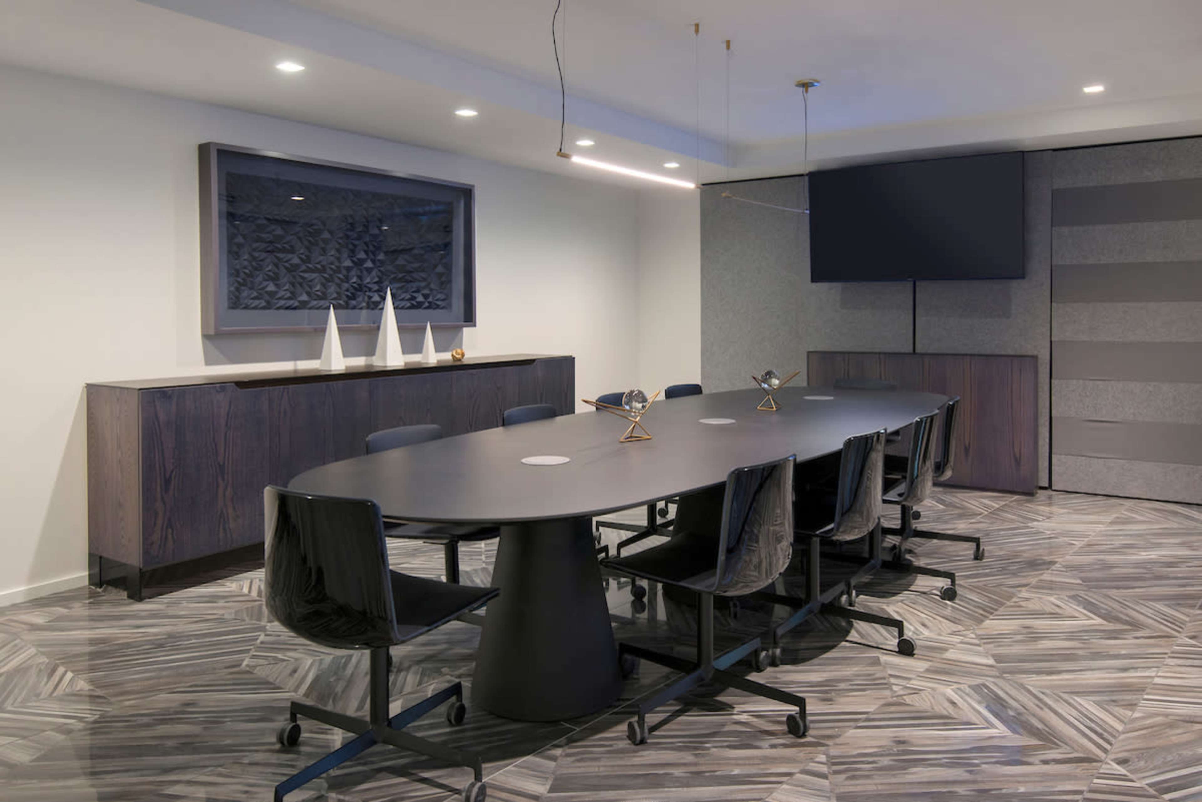 A long, oval conference table is surrounded by rolling black chairs in a modern meeting room featuring a large screen and decorative wall art.