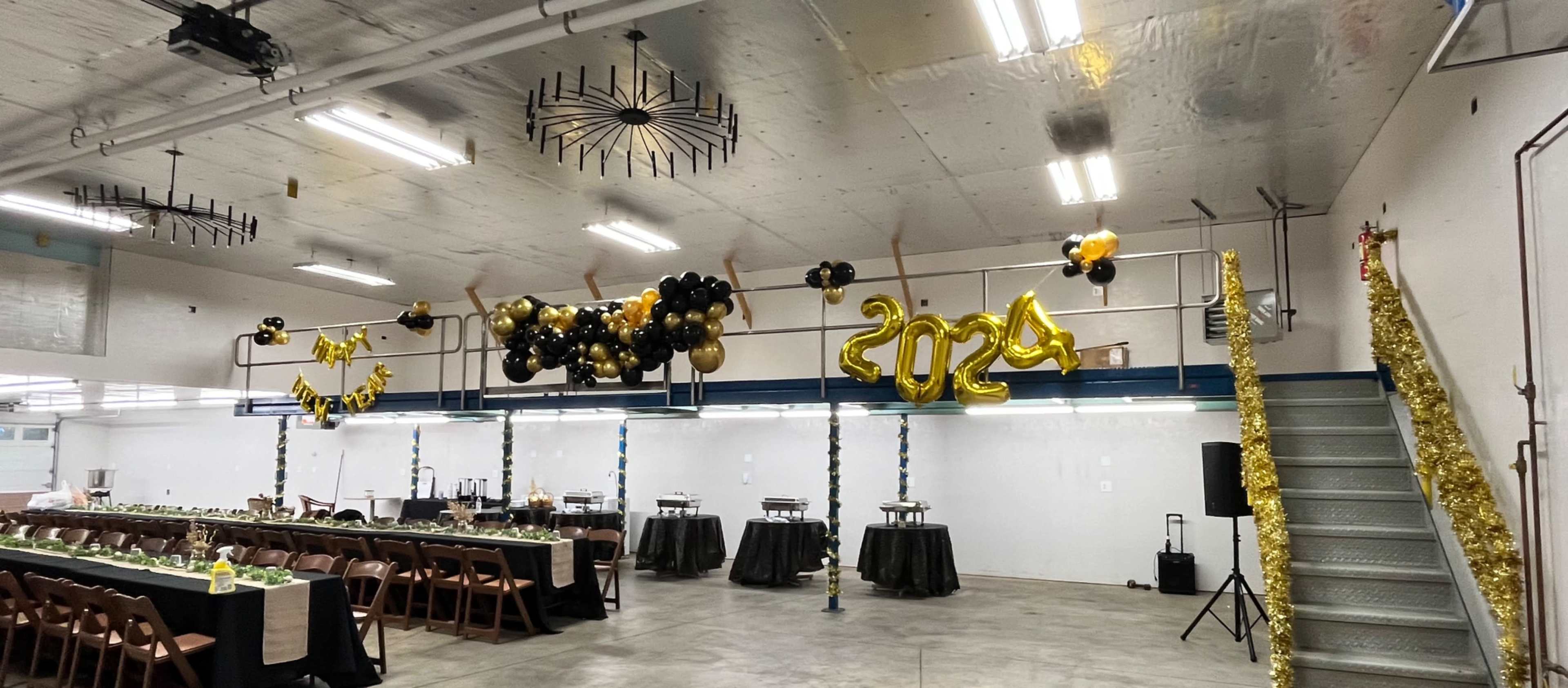 The image shows a decorated event space with black and gold balloons, a long dining table set for a celebration, and a festive “2024” banner hanging above.
