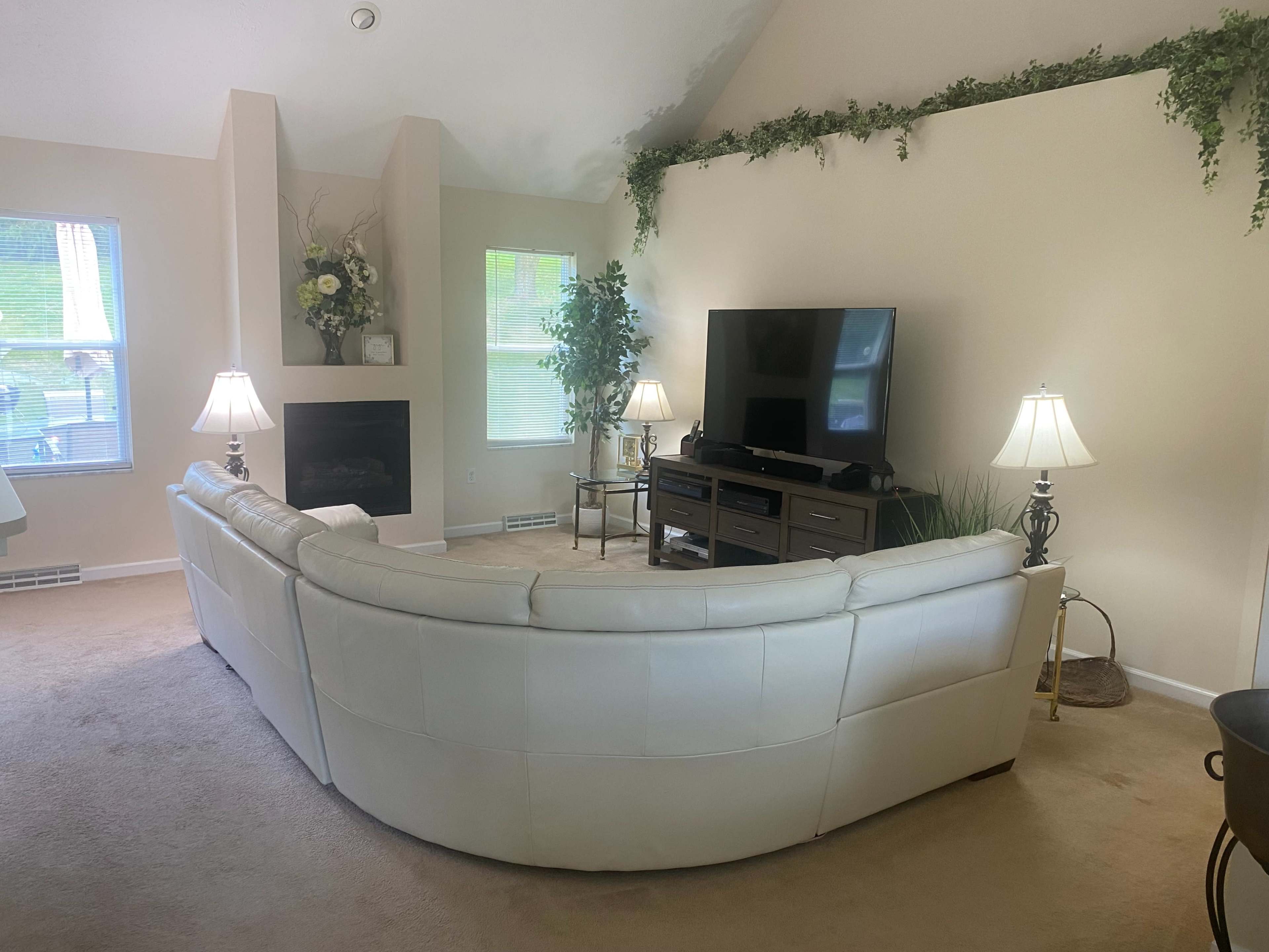 A spacious living room features a large white sectional sofa, a television stand, and decorative plants near the windows.