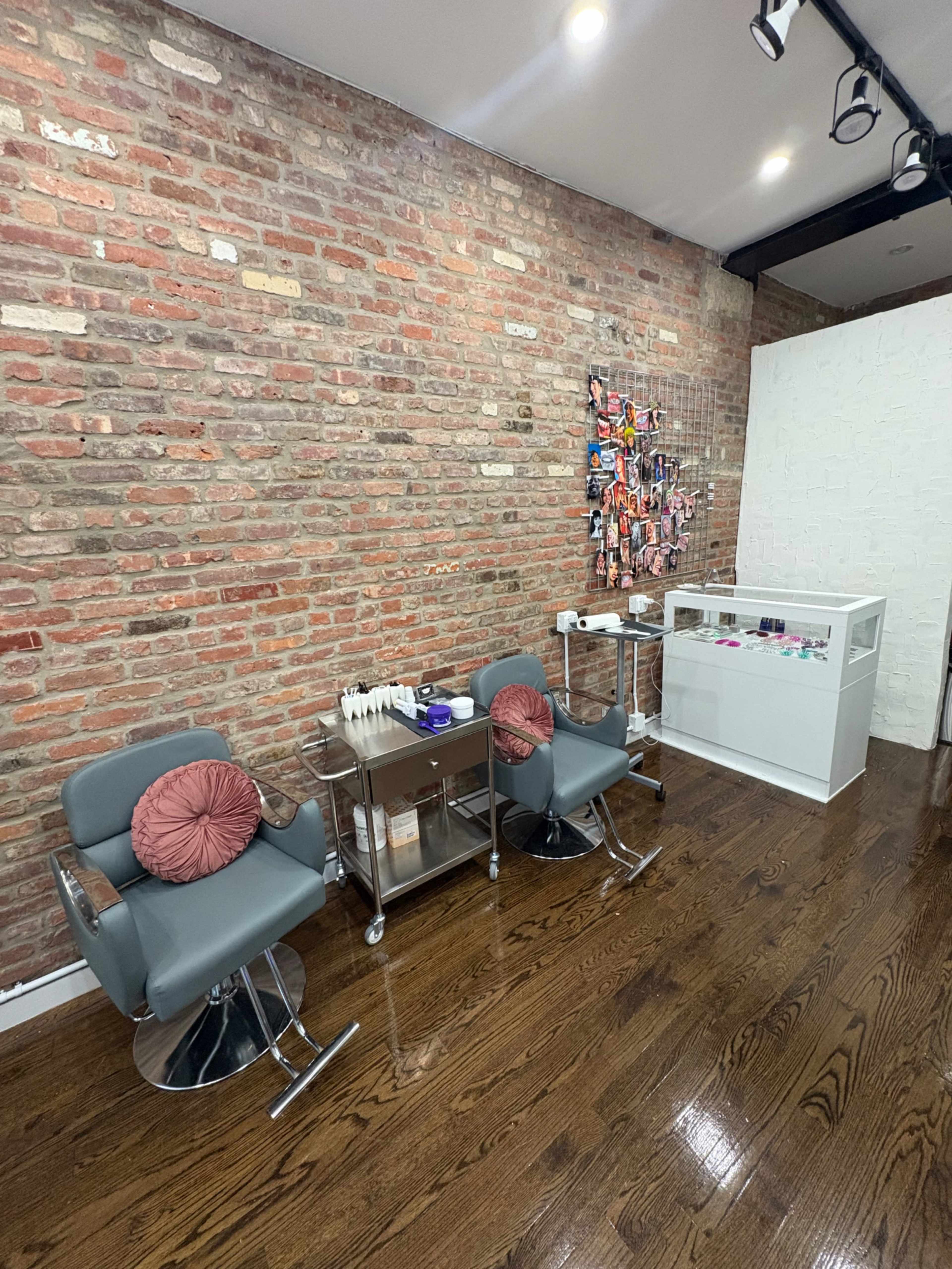 The image shows a modern salon interior featuring two gray chairs, a metal service cart, and a display case against a backdrop of exposed brick walls and polished wooden flooring.