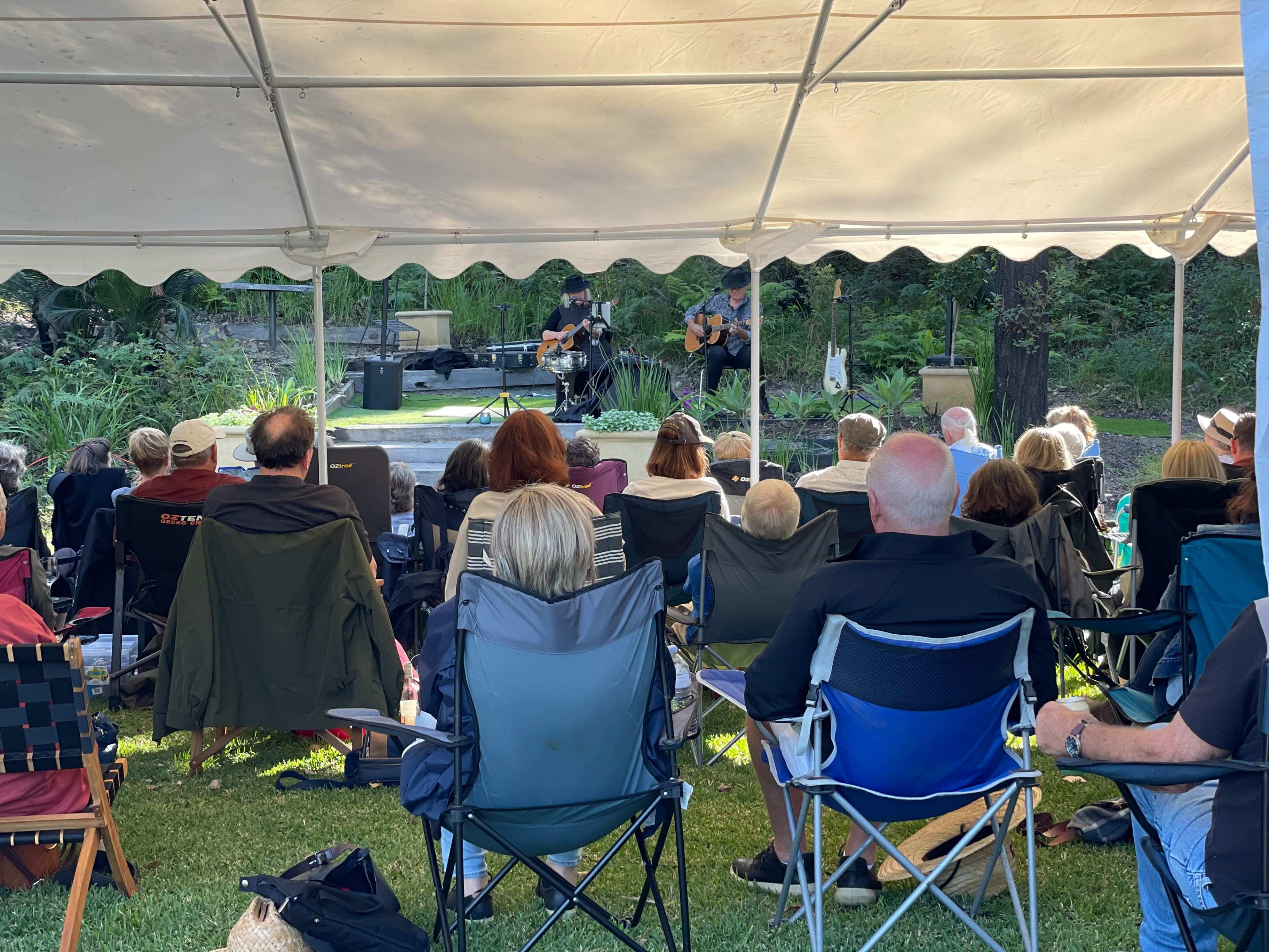 A crowd of seated individuals is gathered under a tent, listening to two musicians performing on a stage.