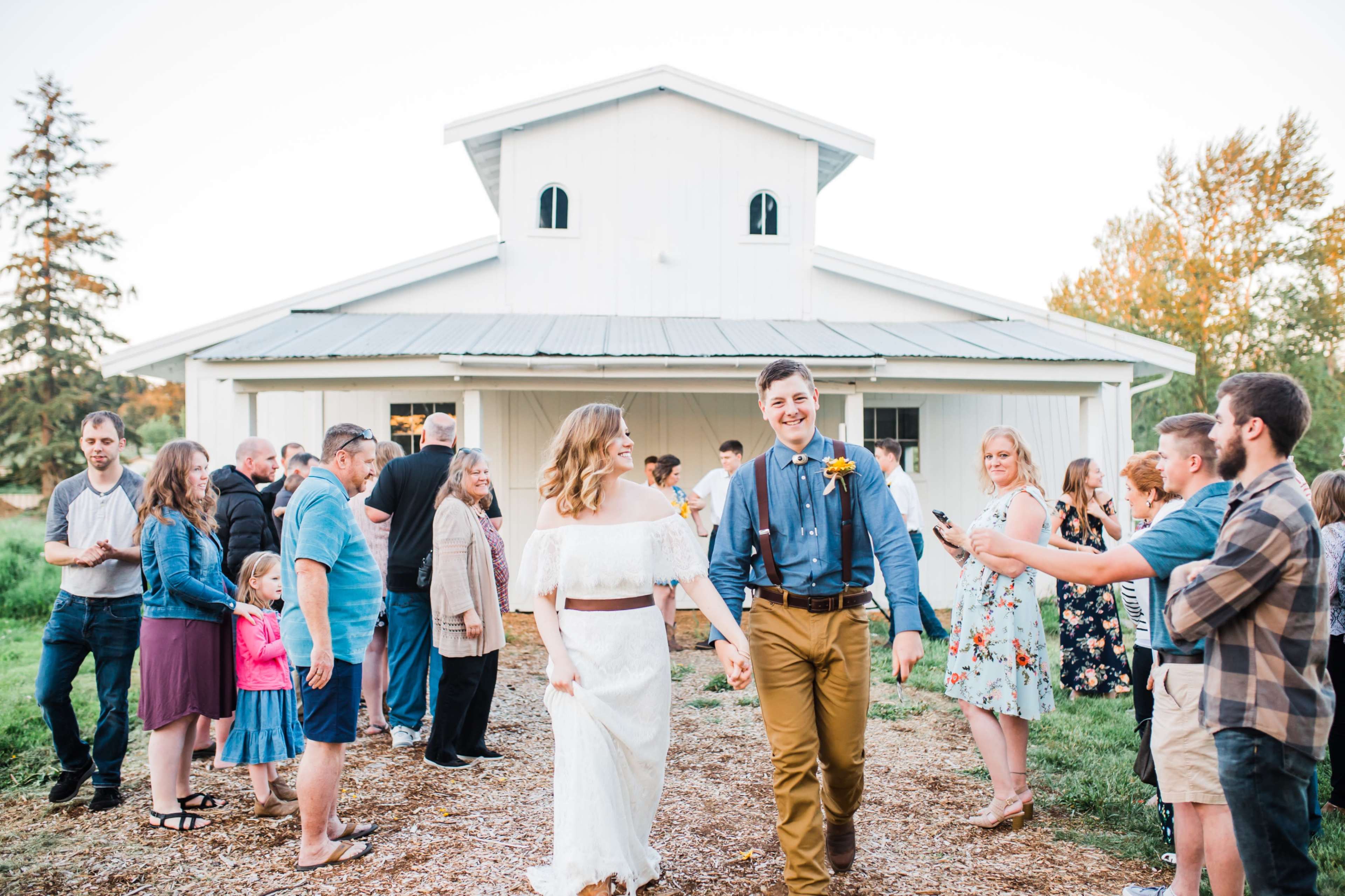 A bride and groom walk hand in hand through a crowd of guests outside a barn during their wedding celebration.