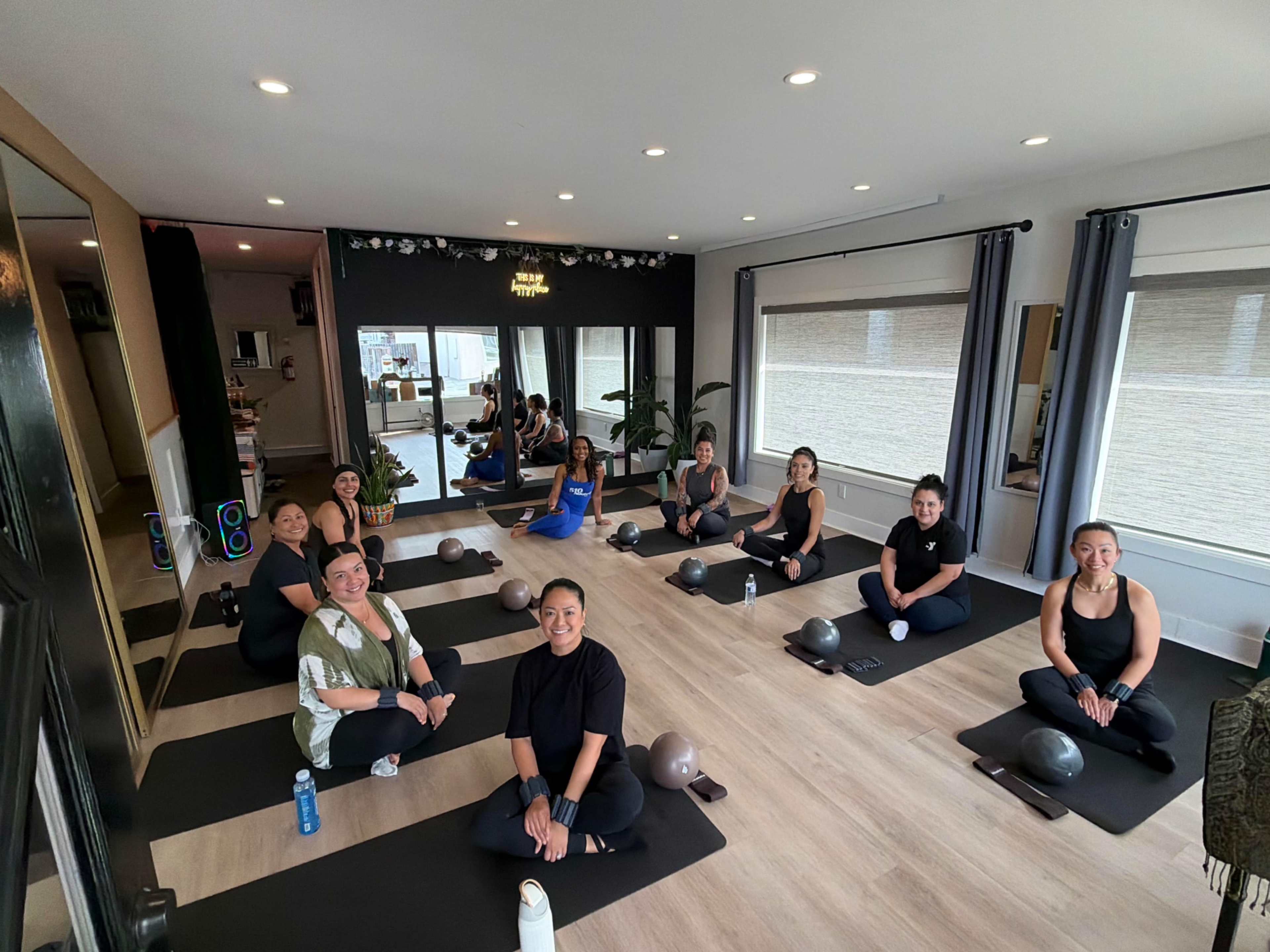A group of nine women is seated on yoga mats in a well-lit studio, preparing for a fitness class.