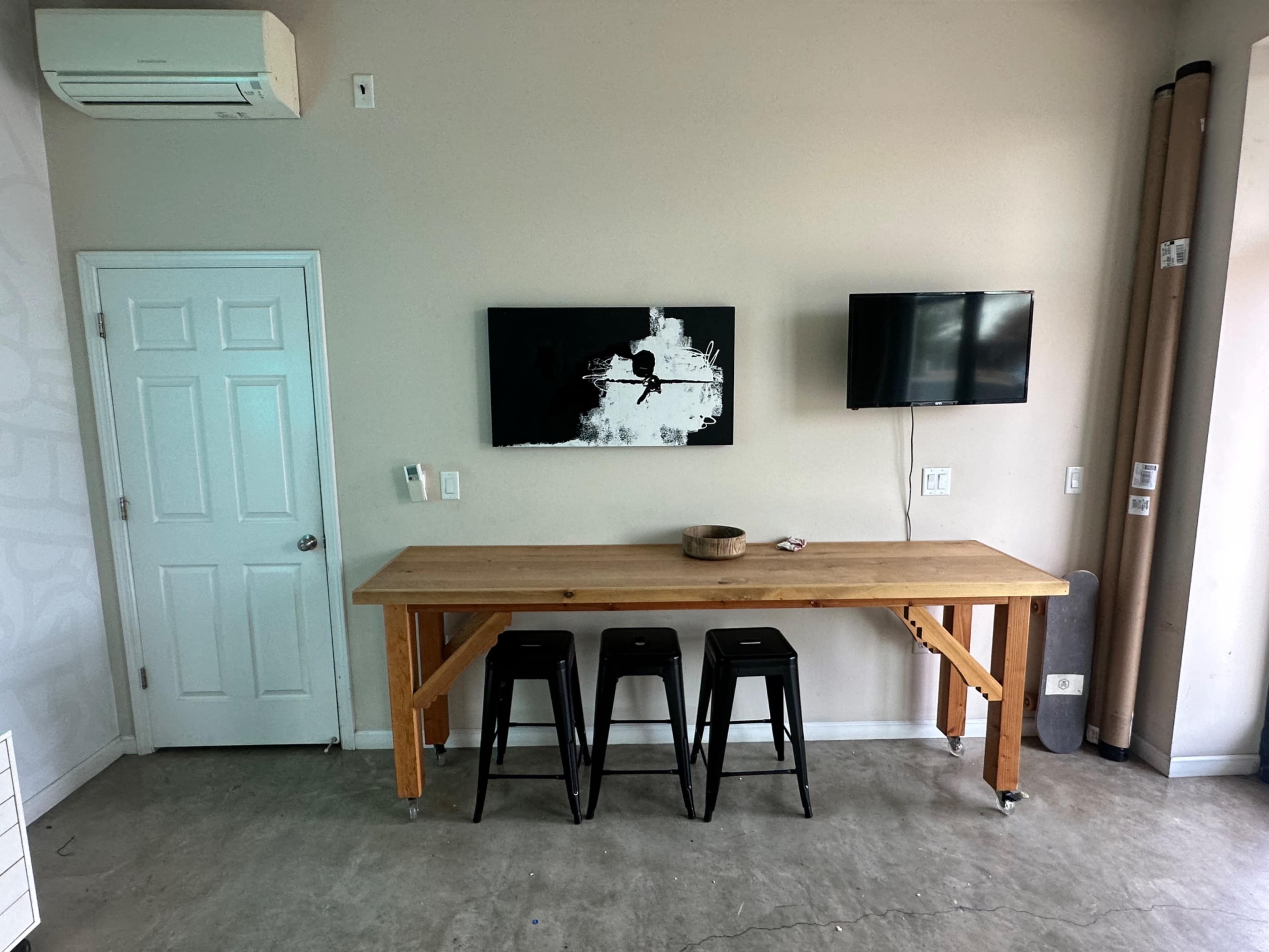 A wooden table with three black stools stands against a wall, featuring two televisions and a piece of artwork above it.