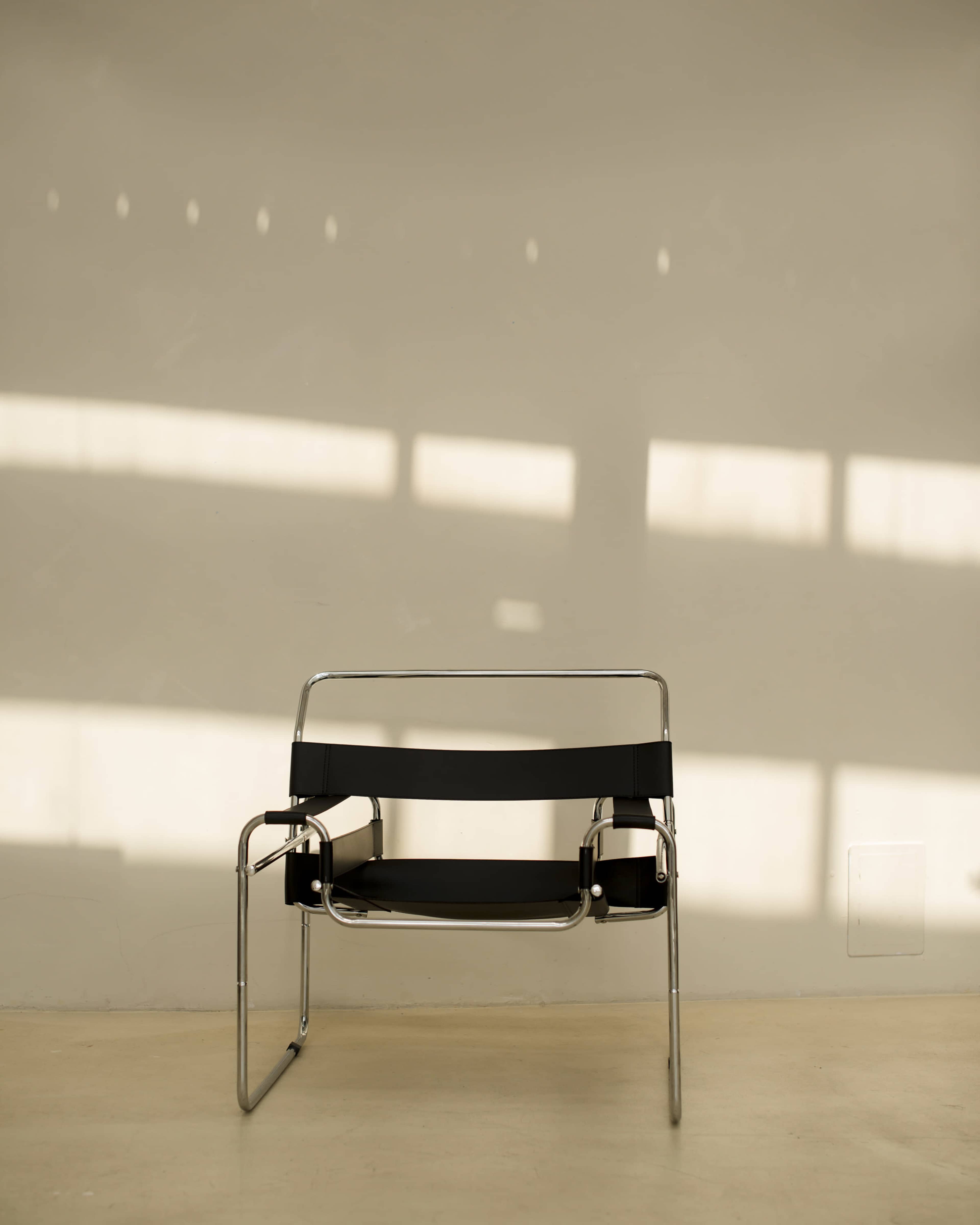 A black chair with a chrome frame sits against a light-colored wall, casting shadows from a nearby window.