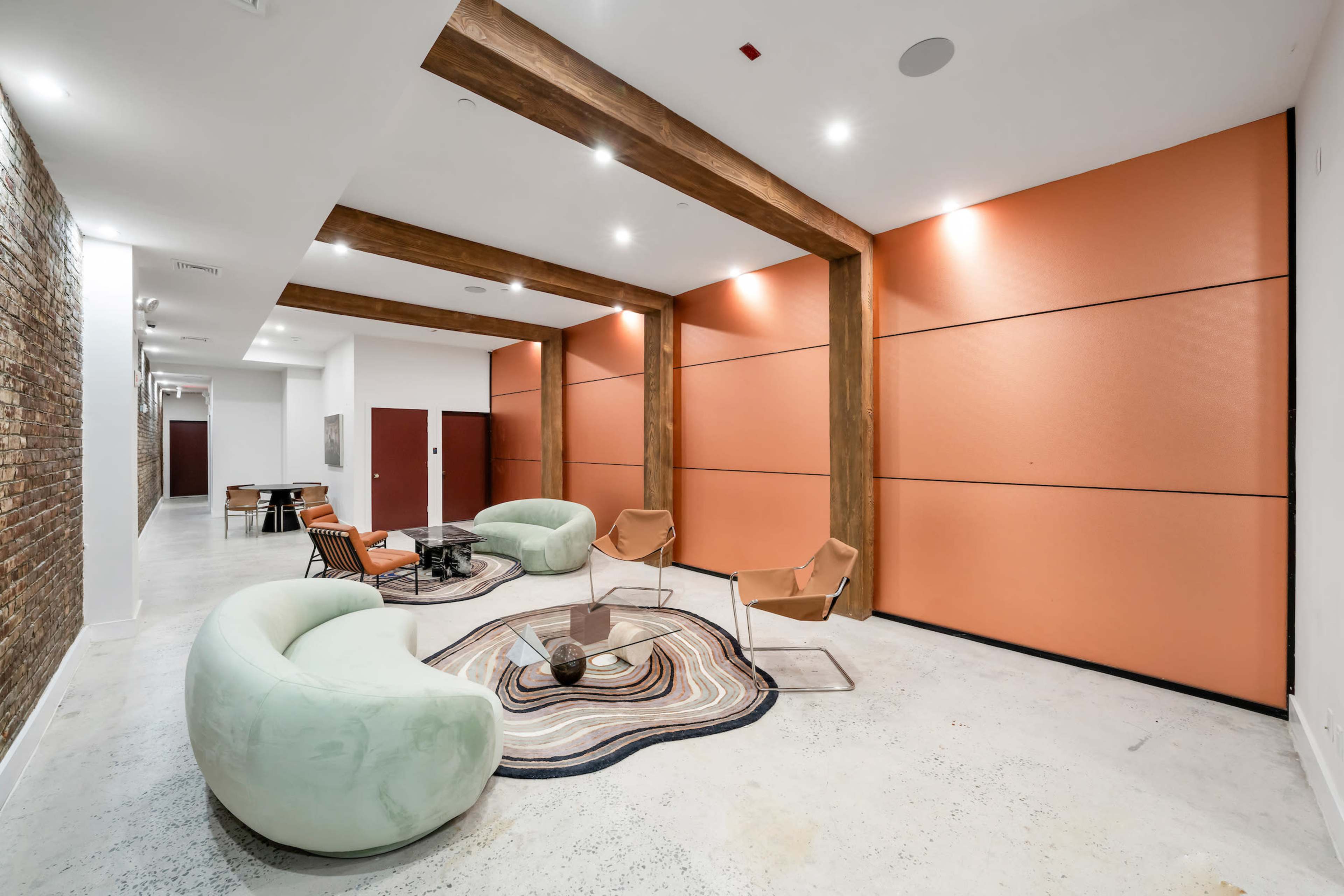 A modern lounge area with two green couches, two tan chairs, a round coffee table, and orange walls accented by wooden beams.
