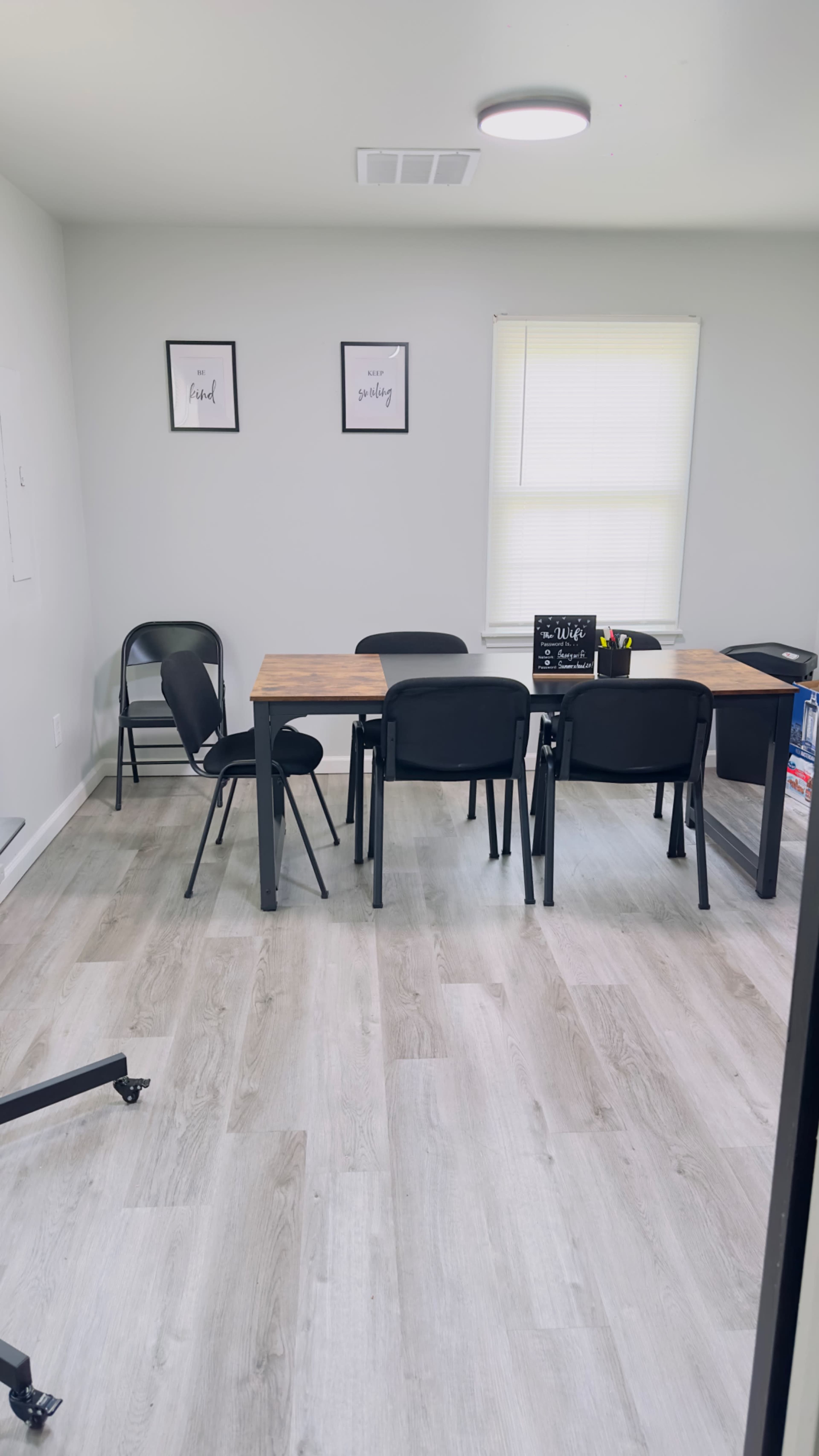 A simple meeting room features a wooden table surrounded by black chairs, with framed artwork on the walls and a window letting in natural light.