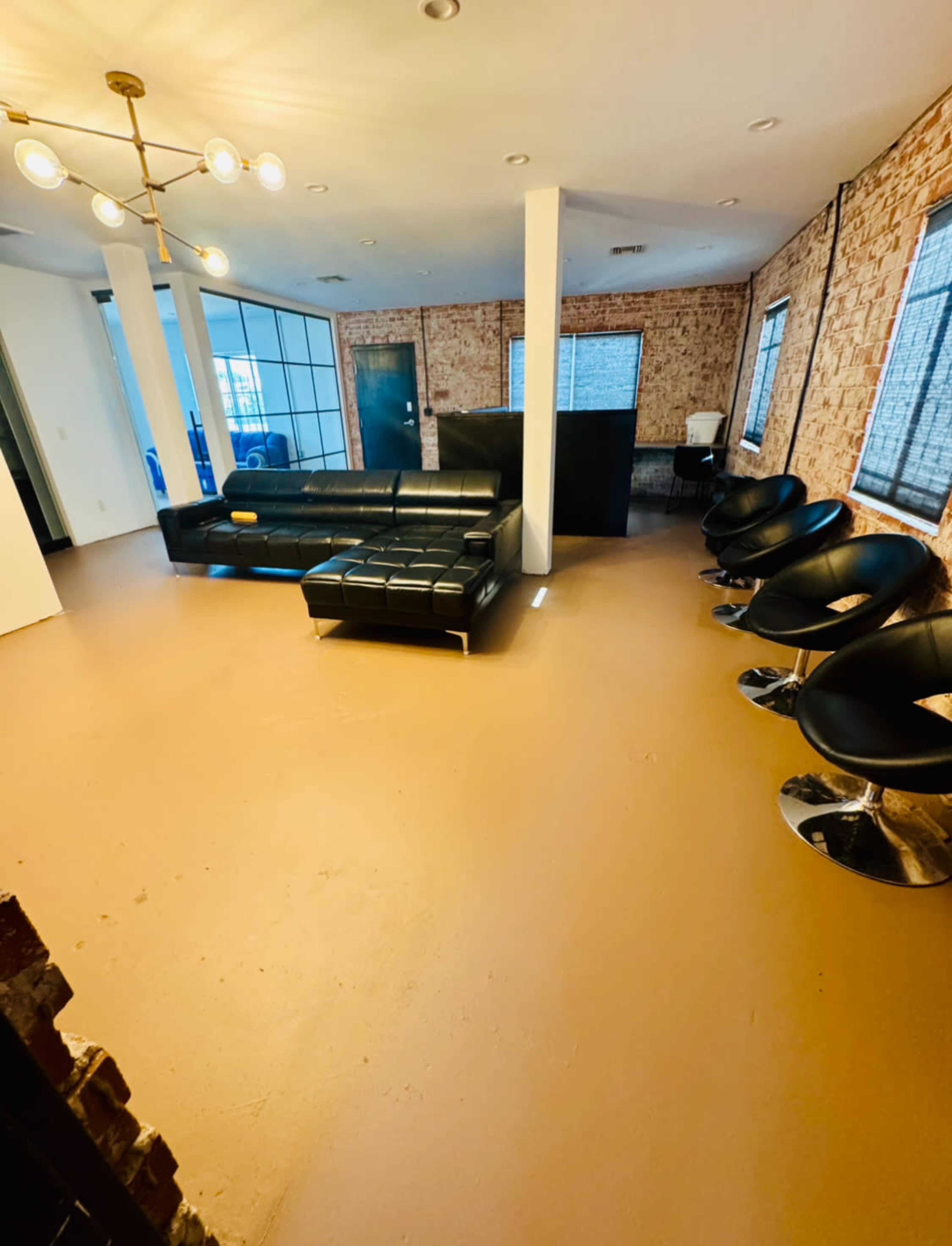 The image shows a spacious, modern room with a black sectional sofa, several black swivel chairs, and exposed brick walls.