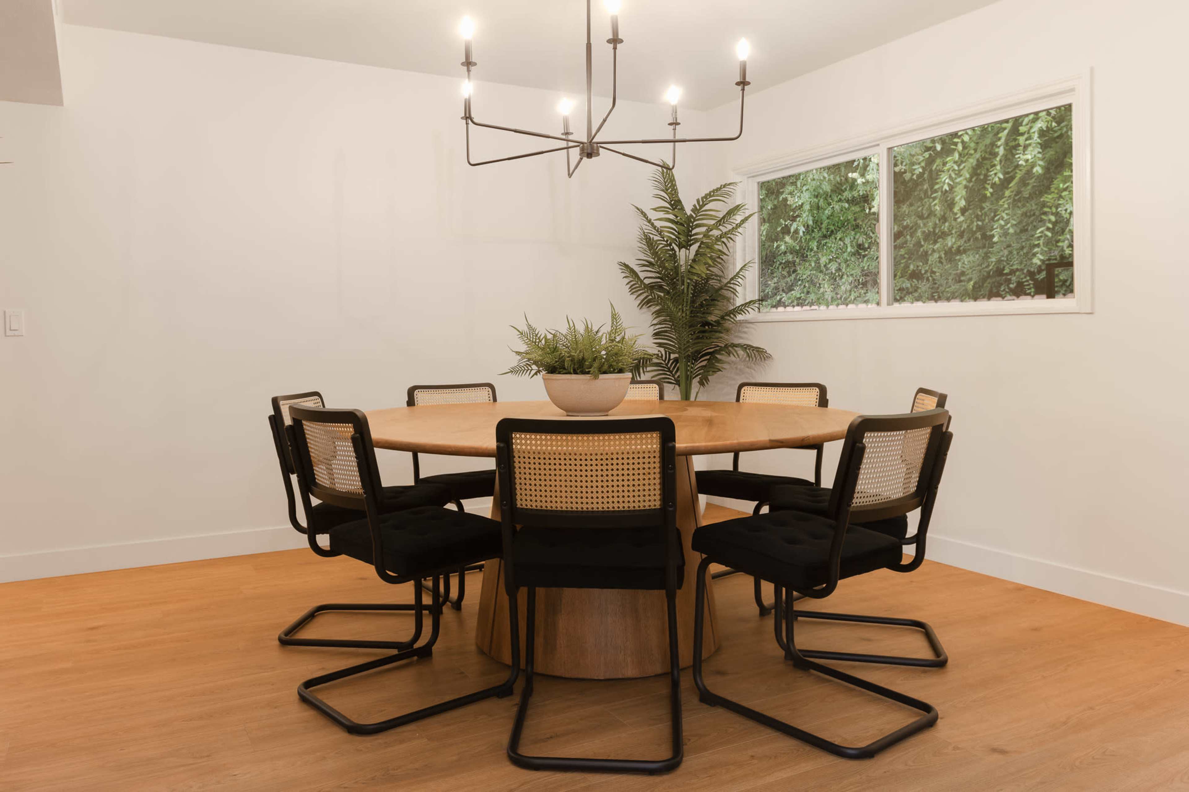 A round dining table is surrounded by six chairs in a well-lit room with a window and a potted plant.