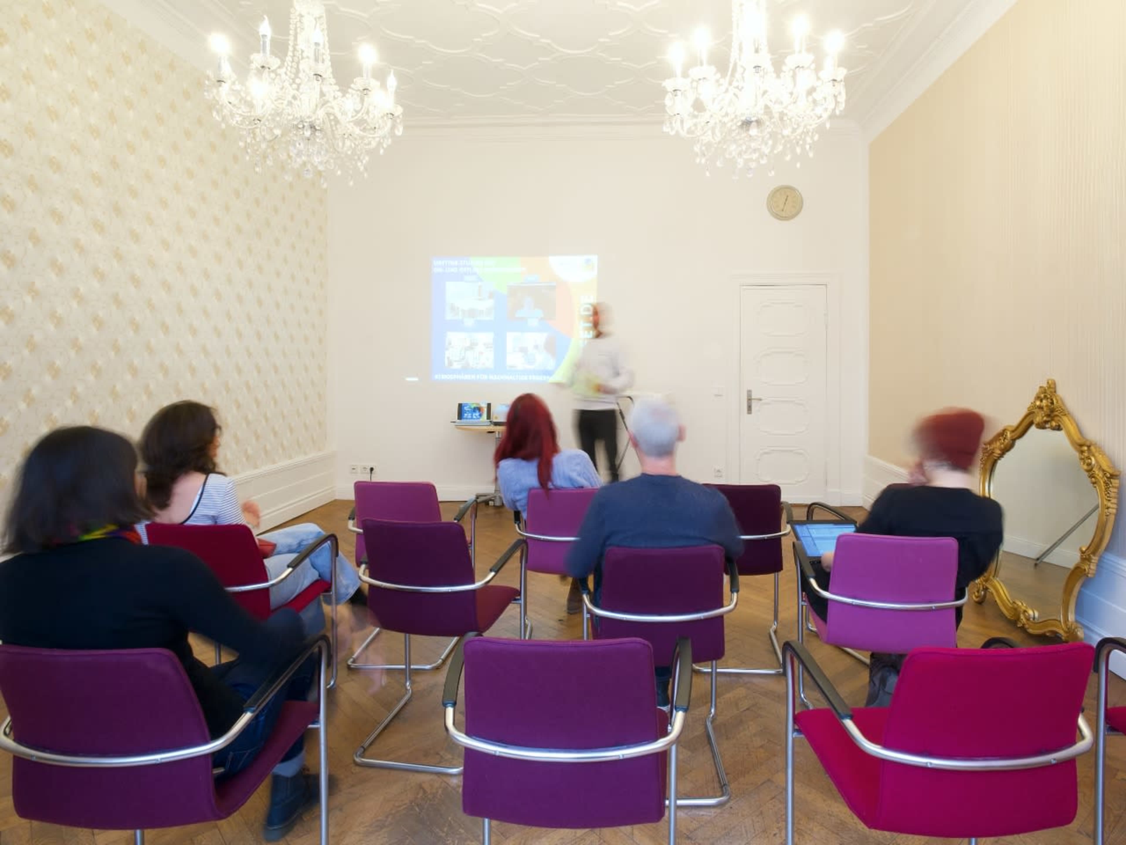 A group of people sits in chairs facing a projector screen in a well-lit room with chandeliers and ornate decor.
