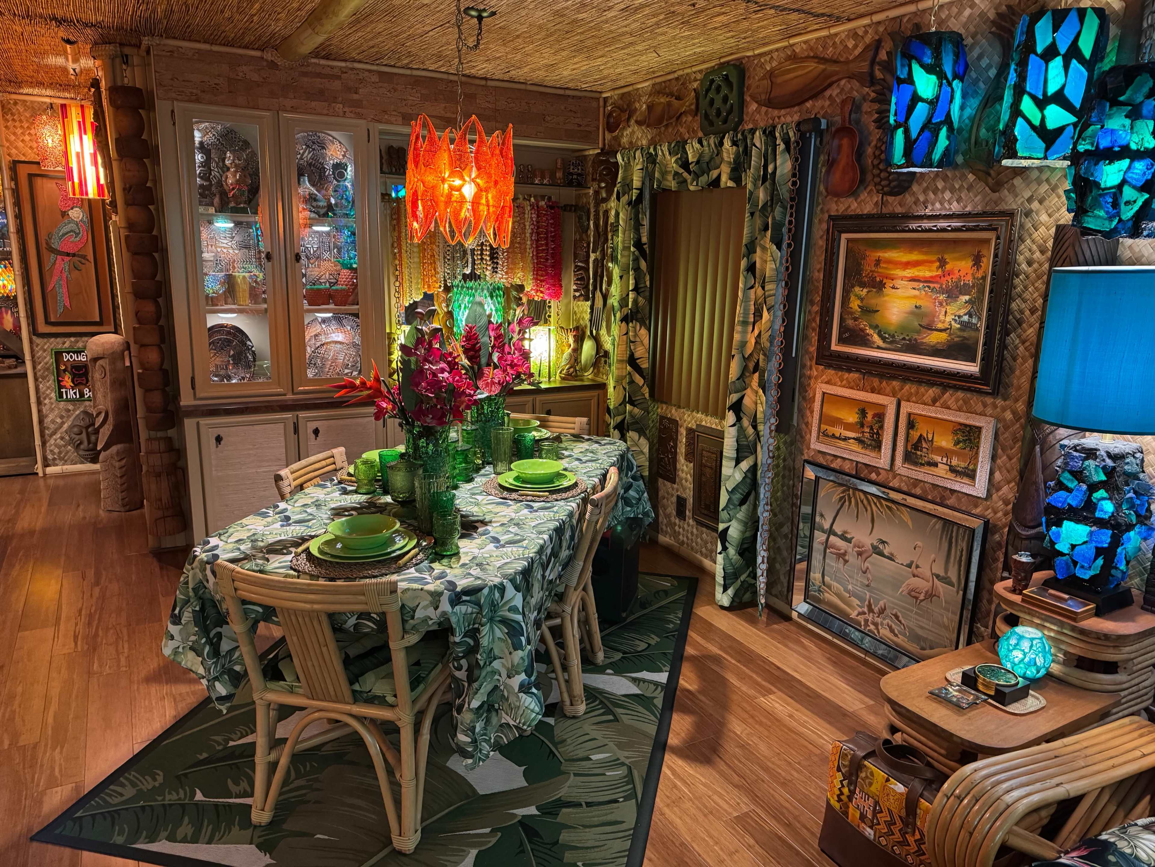 The image shows a tropical-themed dining room with a table set for a meal, featuring floral tablecloth and green dishware, surrounded by colorful decor and artwork.