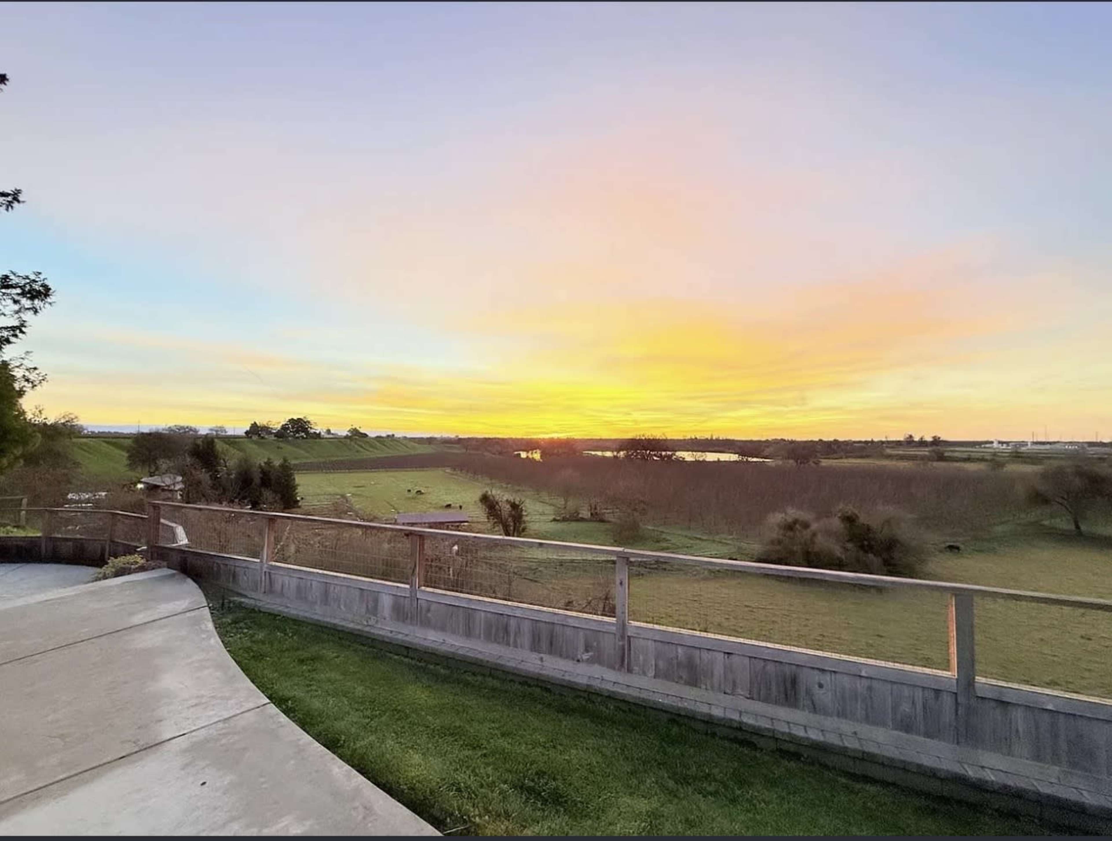 The image shows a landscape with a foreground of a concrete path and wooden fence, leading to rolling green fields and a sunset sky with vibrant colors.