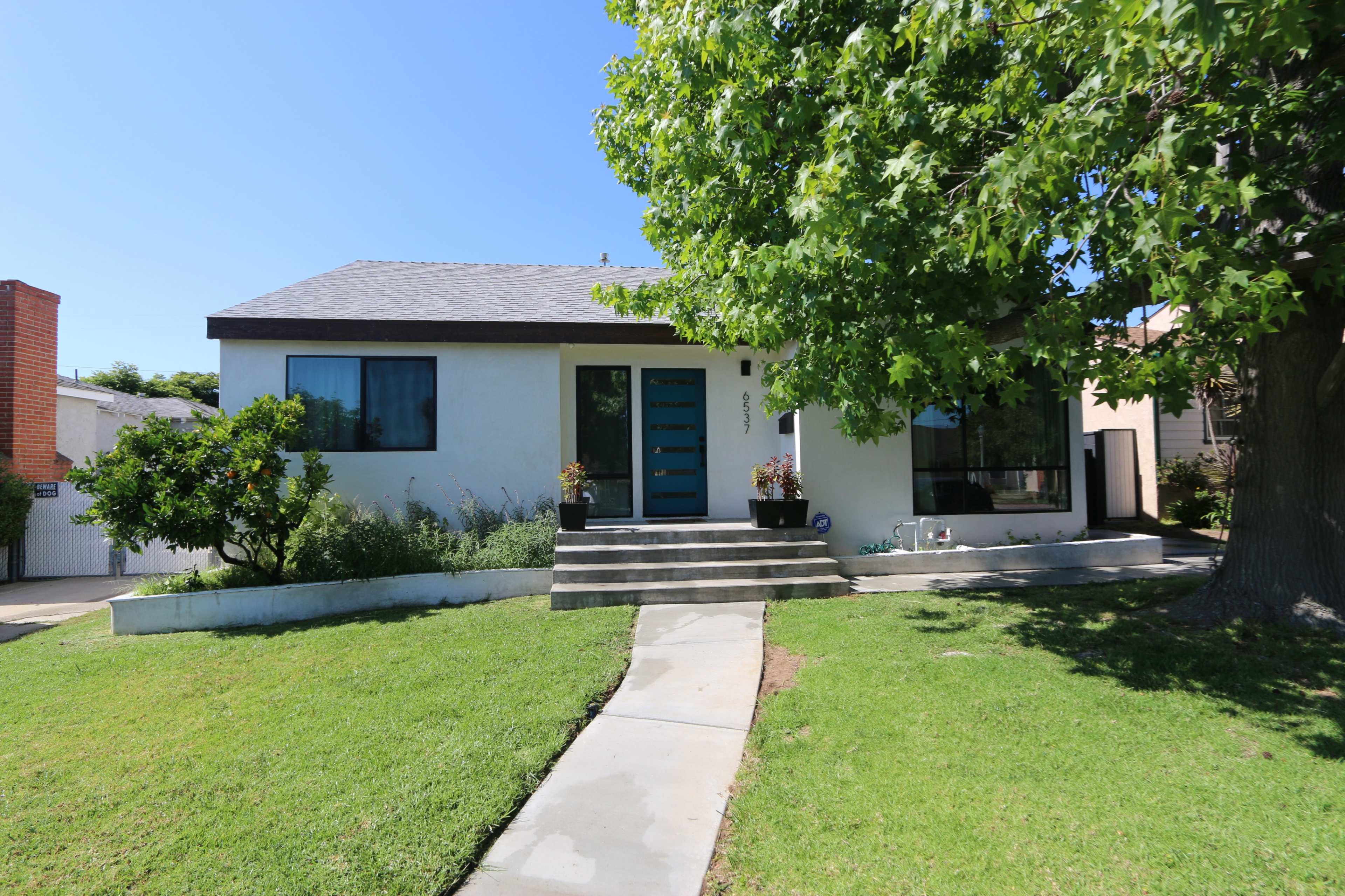 A single-story house with a flat roof is flanked by a well-maintained lawn and a large tree, featuring a front walkway leading to a modern double door entrance.