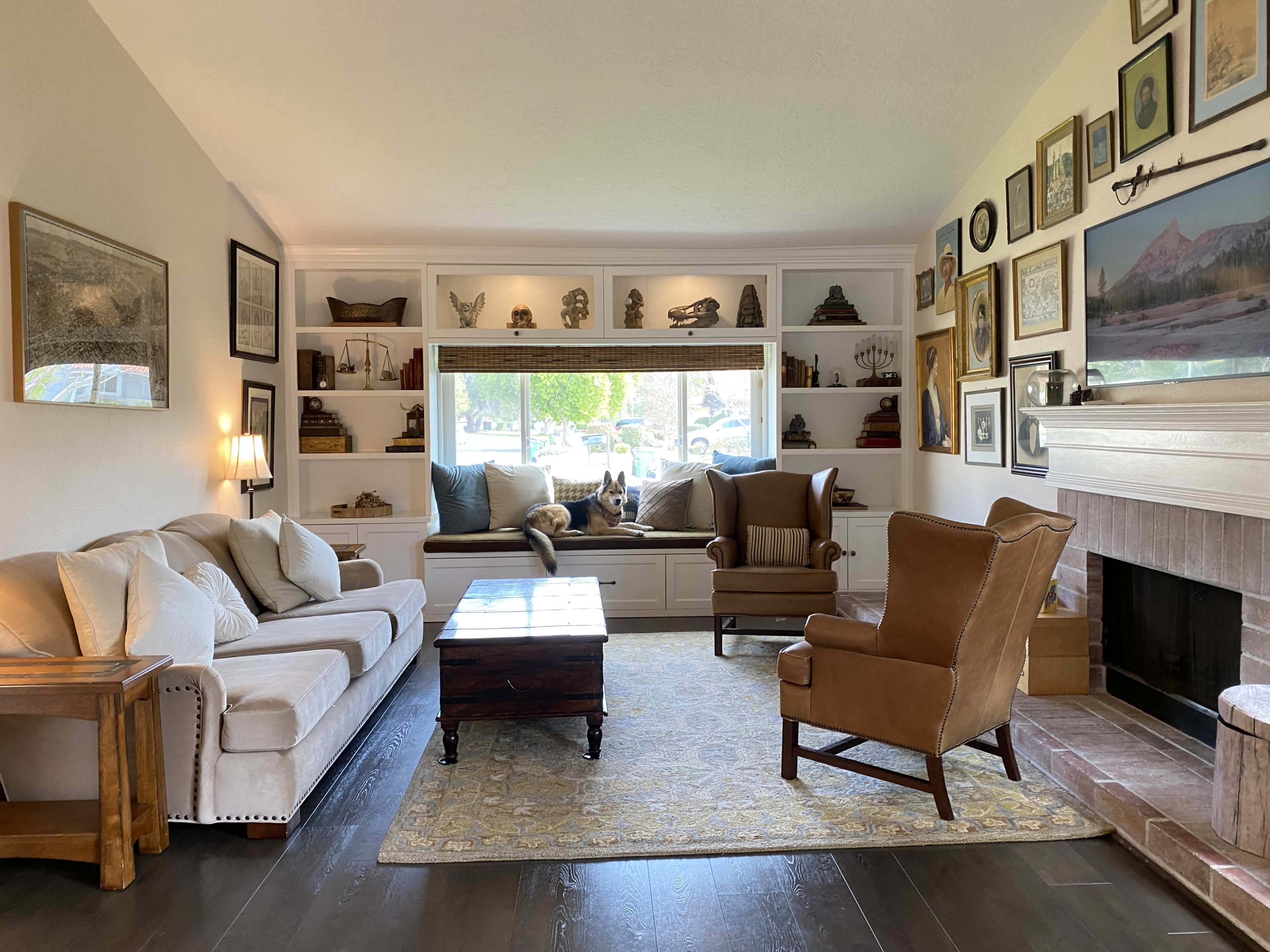 A living room features a beige sofa, a wooden coffee table, and an armchair, with built-in shelves displaying various decorative items and a window seat in the background.