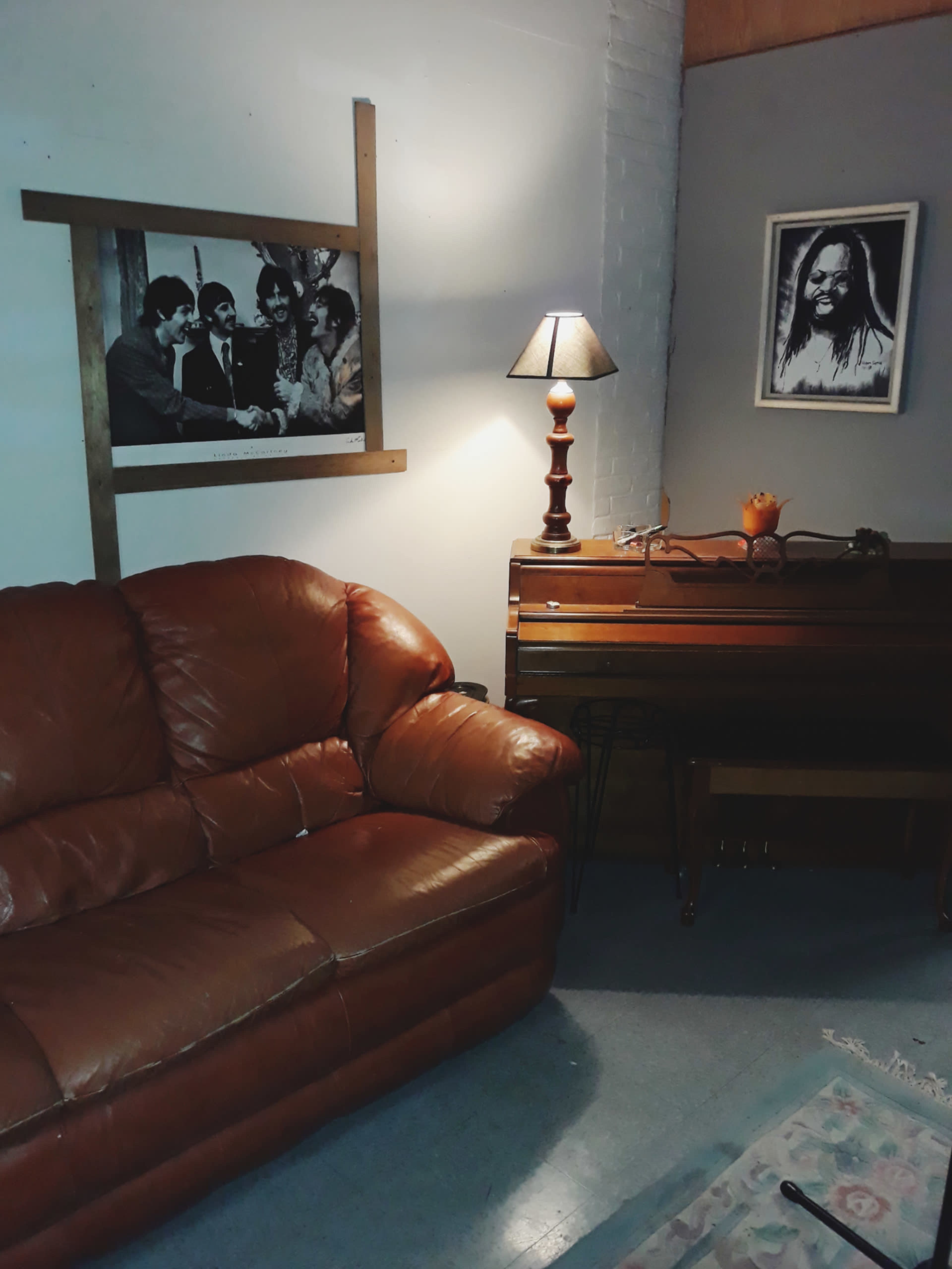 A modest living room with a brown leather couch, a lamp, a wooden piano, and framed photographs on the walls.