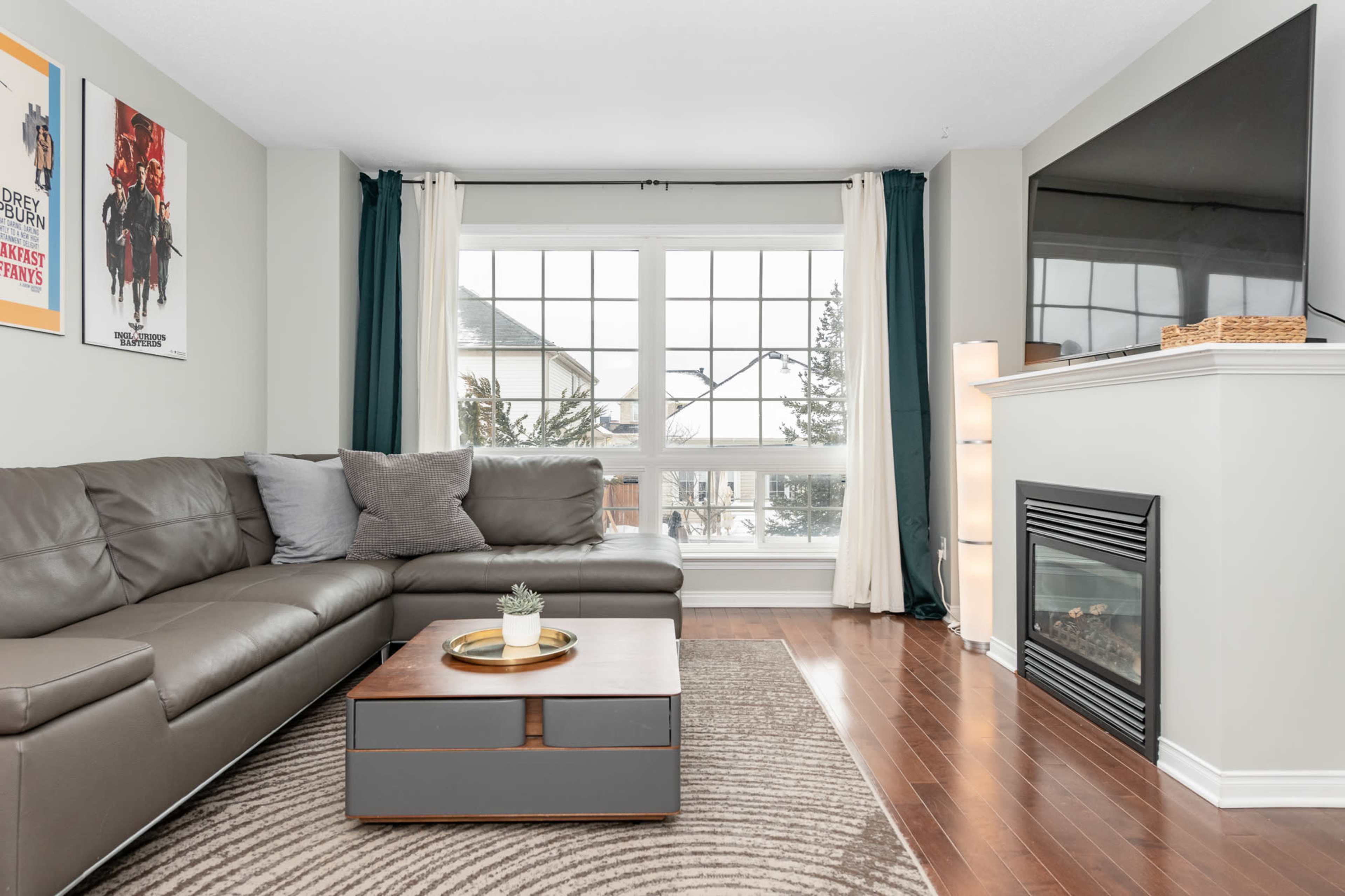 A living room with a gray sectional sofa, a wooden coffee table, and large windows allowing natural light, adorned with curtains.