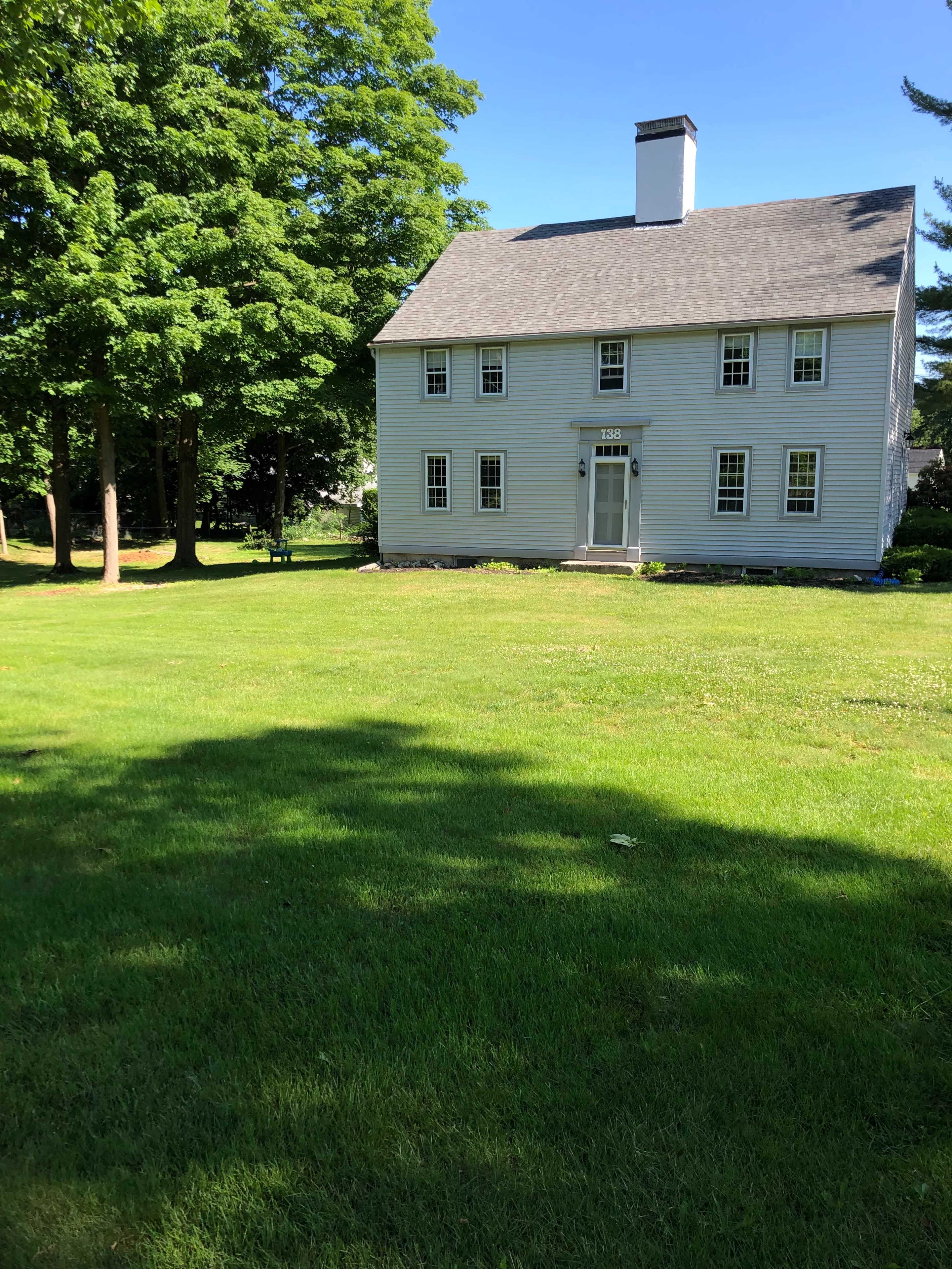 A two-story gray house with white trim stands on a well-maintained lawn surrounded by green trees.